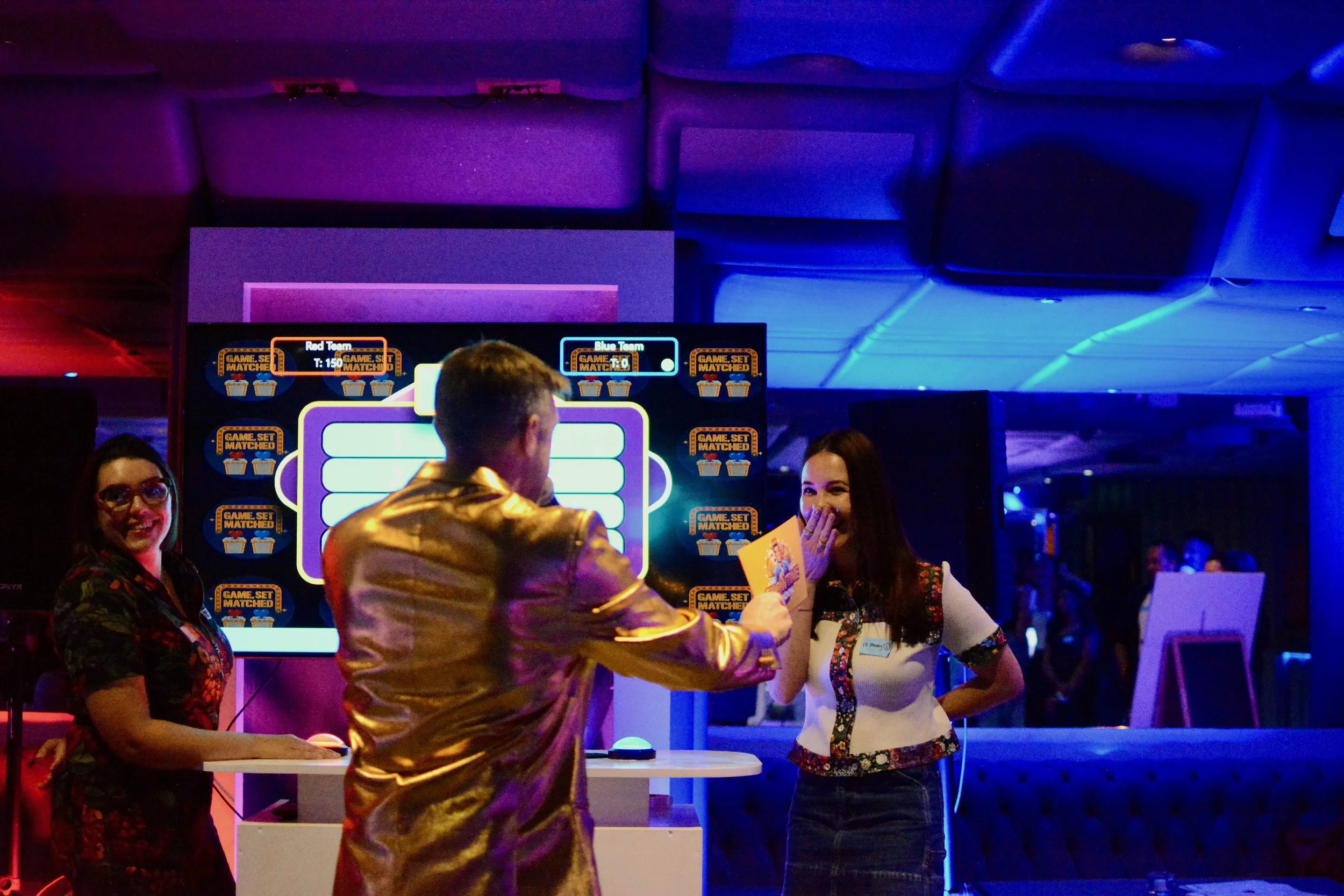 Three people at a game show event, with one man in a shiny gold jacket handing a card to a woman in a decorative top, while another woman looks on. There is a large electronic screen in the background with game show graphics.