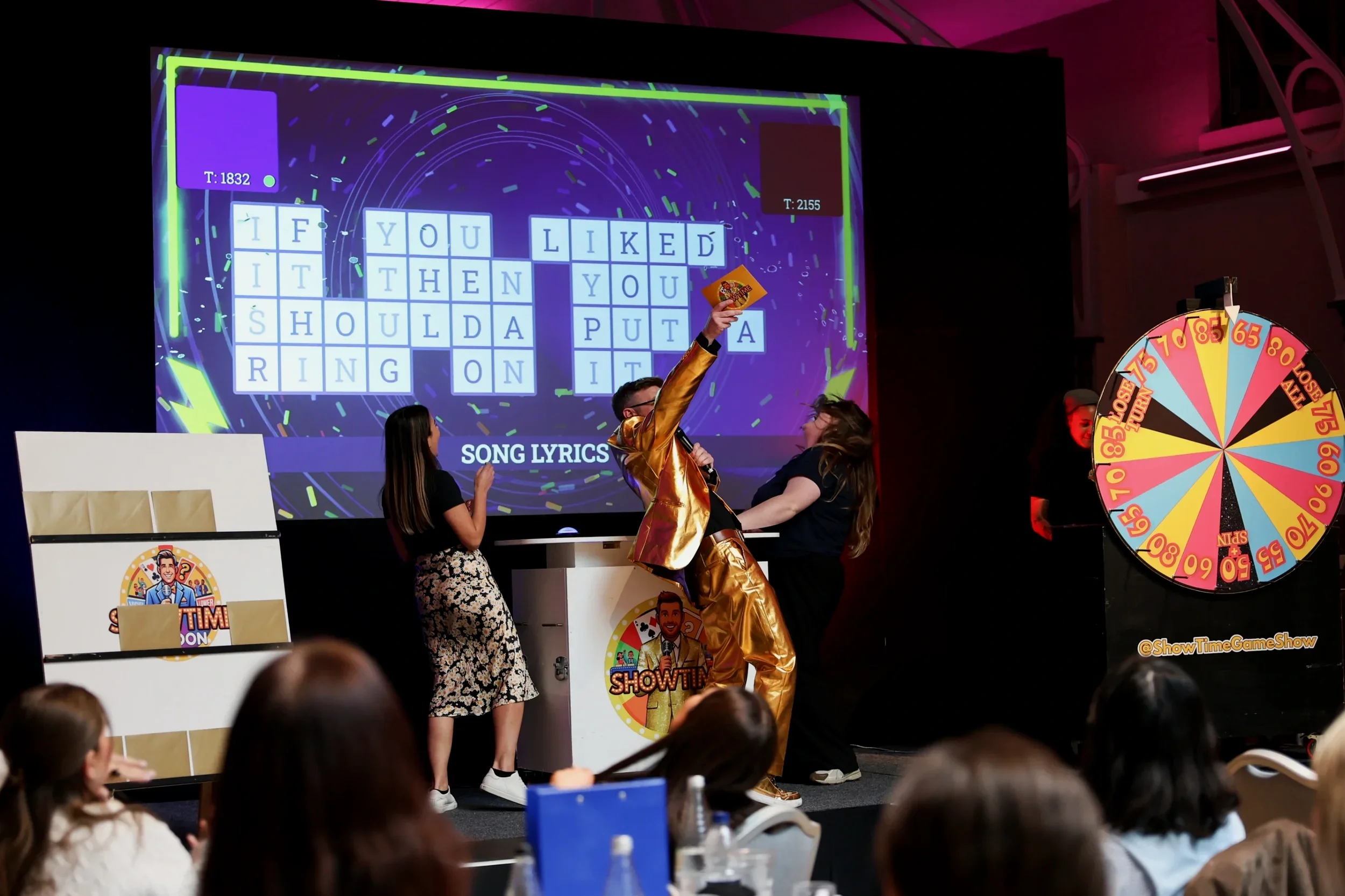 People playing a game show with a large screen displaying song lyrics, a spinning wheel, and a host in a shiny gold jacket.