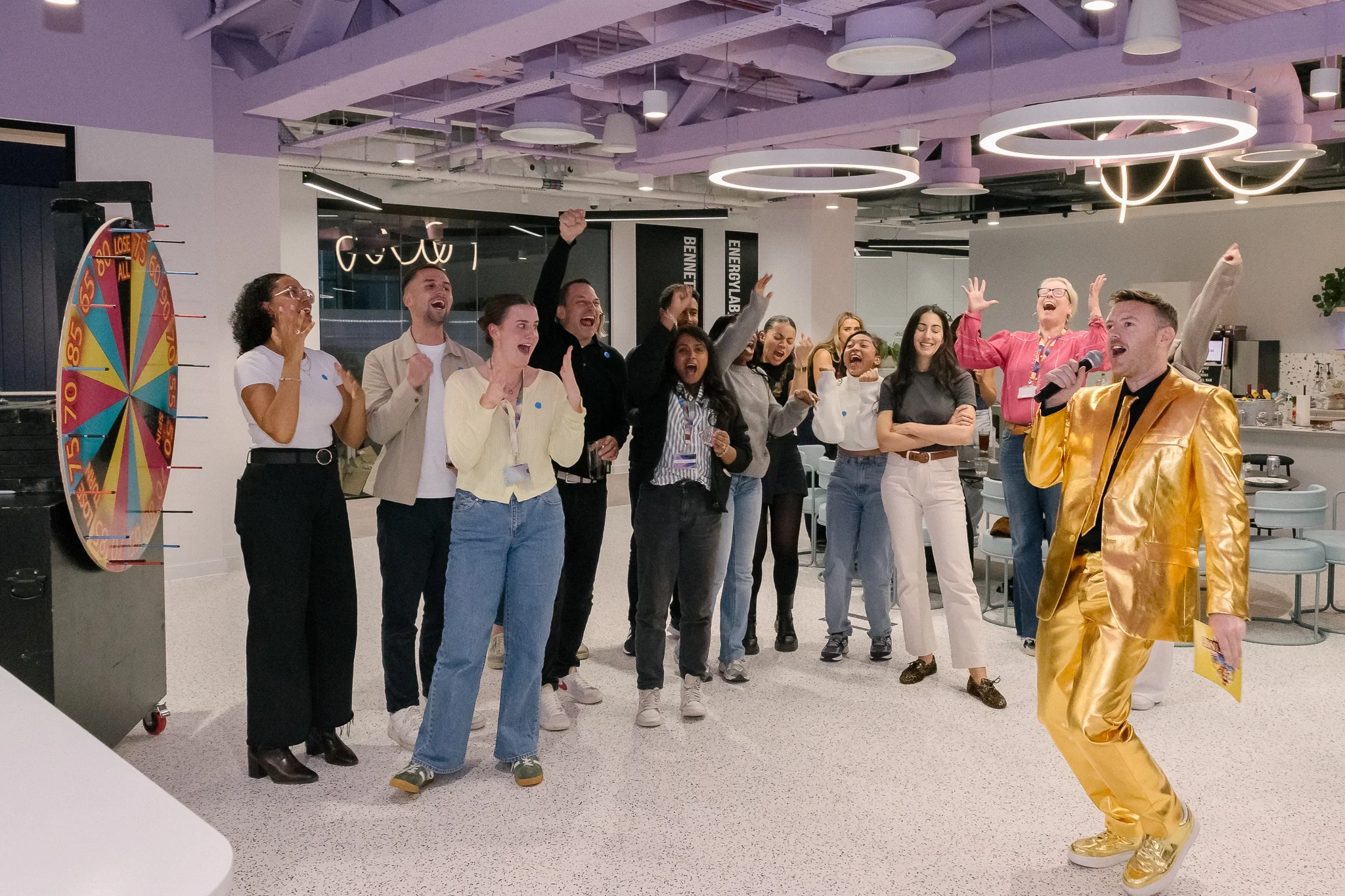 Group of people celebrating, some with hands raised, in a modern office space with colorful lighting and a spinning prize wheel.
