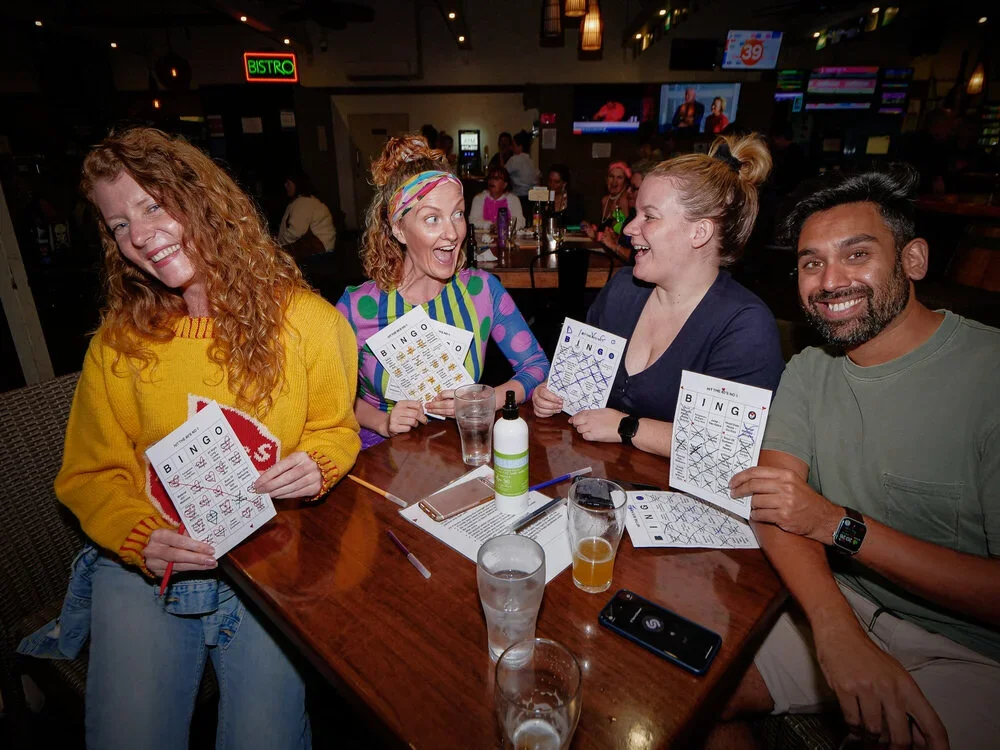 Four friends sitting at a restaurant table playing bingo, smiling and holding bingo cards, with drinks and alcohol on the table.