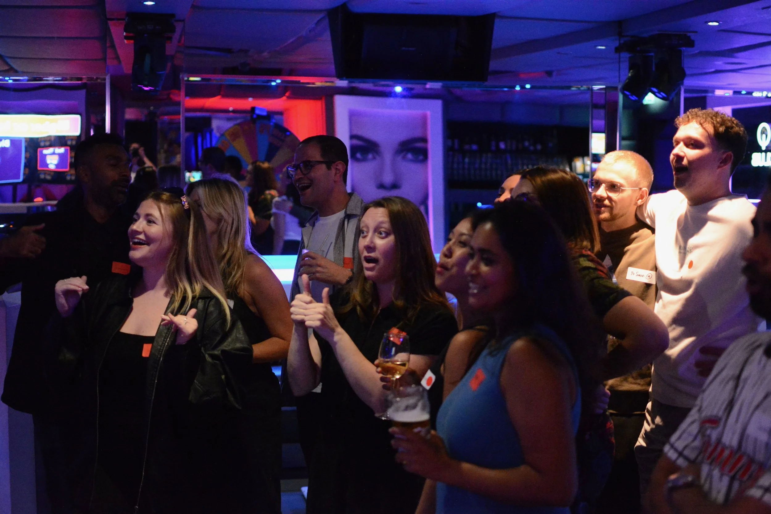 Group of people at a social event, some holding drinks, smiling, and engaging with each other.