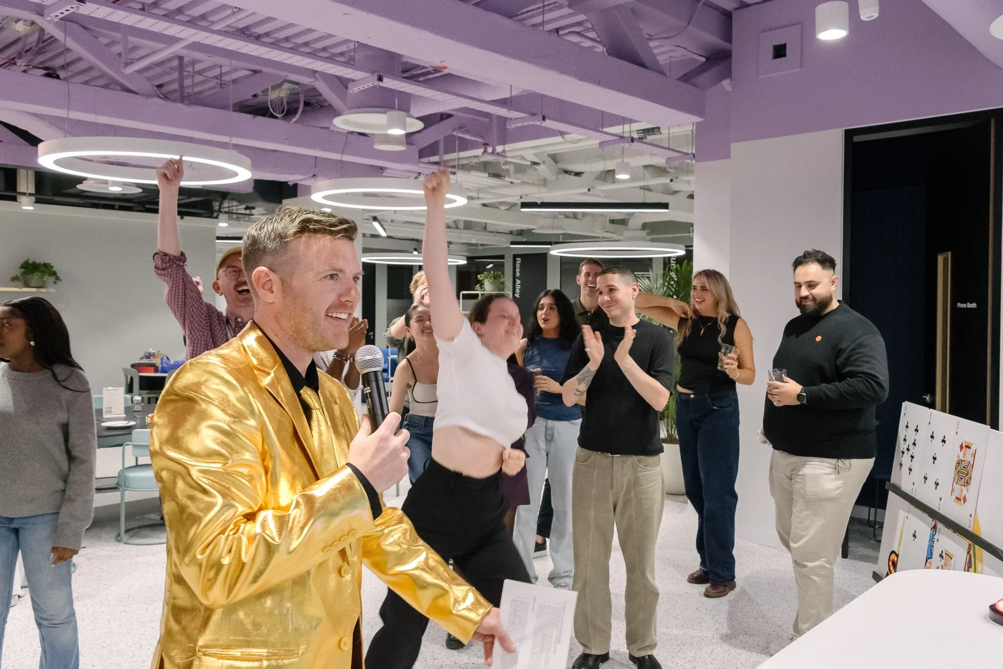 Group of people celebrating indoors, with one man in a shiny gold jacket speaking into a microphone, others clapping and smiling, some holding drinks, festive atmosphere.