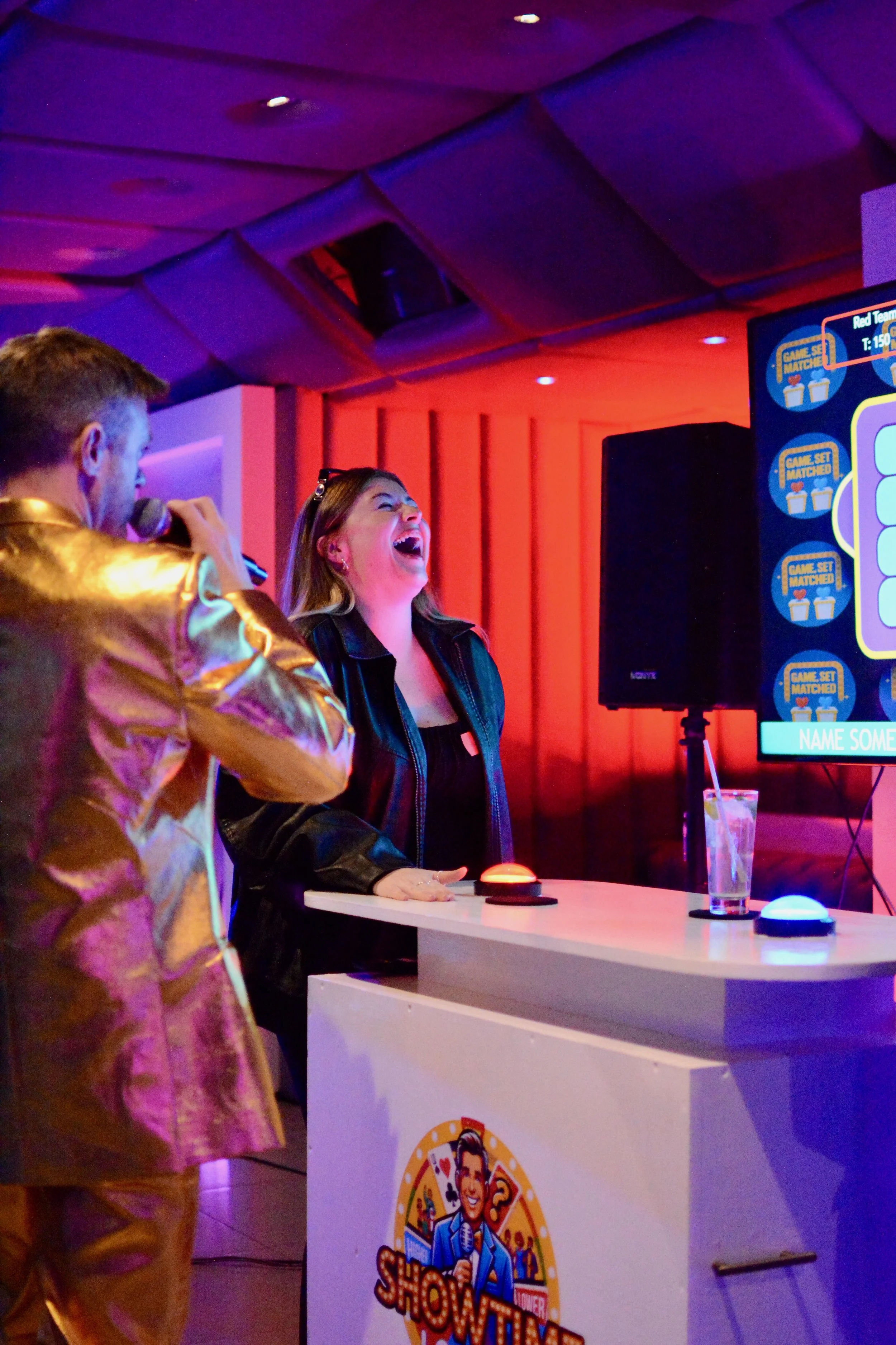 A woman is laughing during a game show, standing at a white podium with a logo, with a man in a shiny gold jacket holding a microphone nearby. There are colorful buttons and a screen with game instructions behind them.