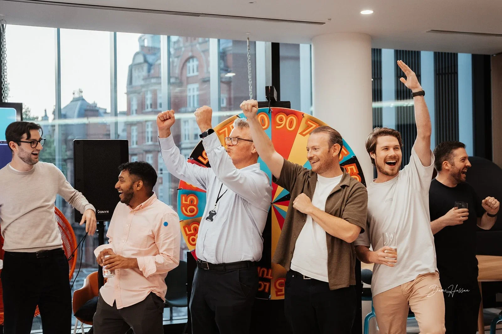 Group of six men celebrating in office with a prize wheel in background
