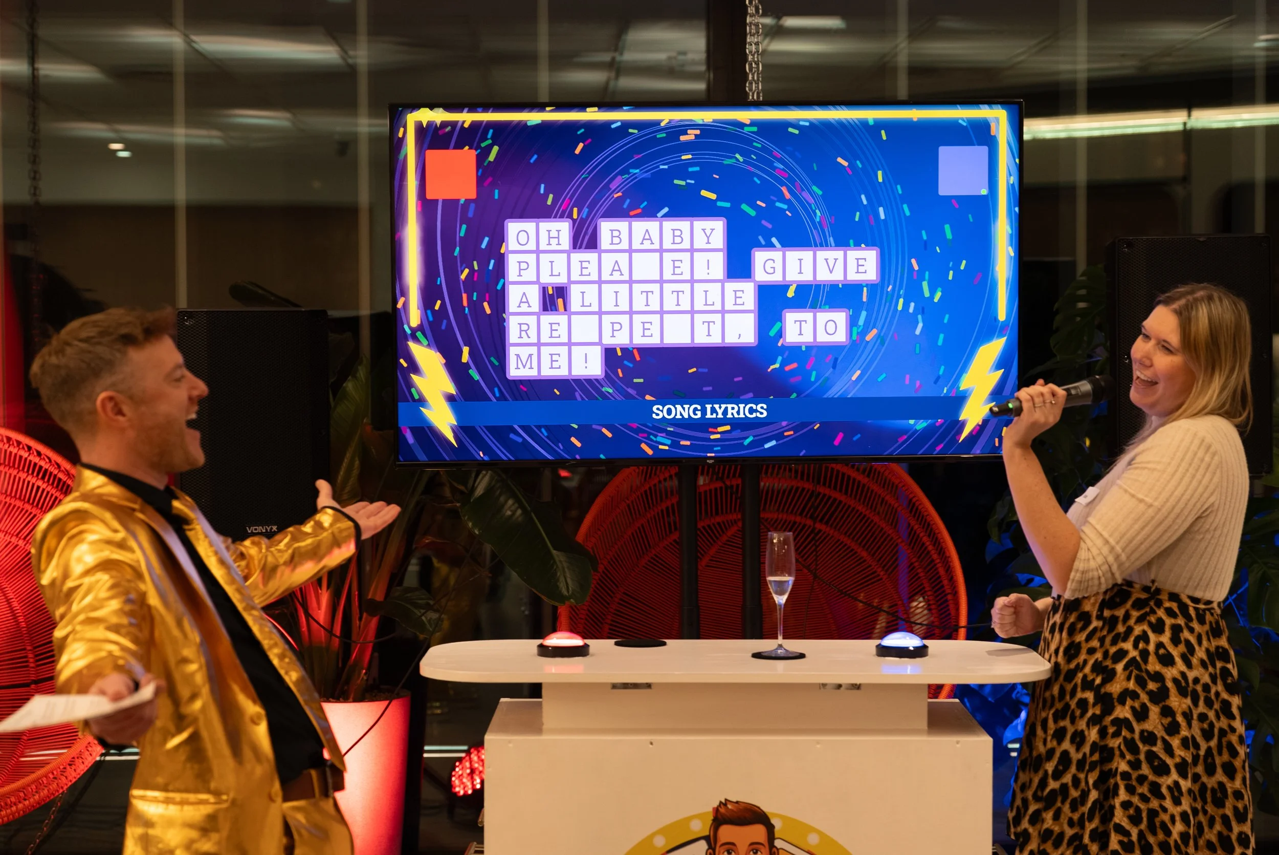 Two people playing a gamesho at a party, with a large scree displaying a word puzzle and song lyrics. The man on the left is wearing a shiny gold jacket, and the woman on the right is holding a microphone, smiling.