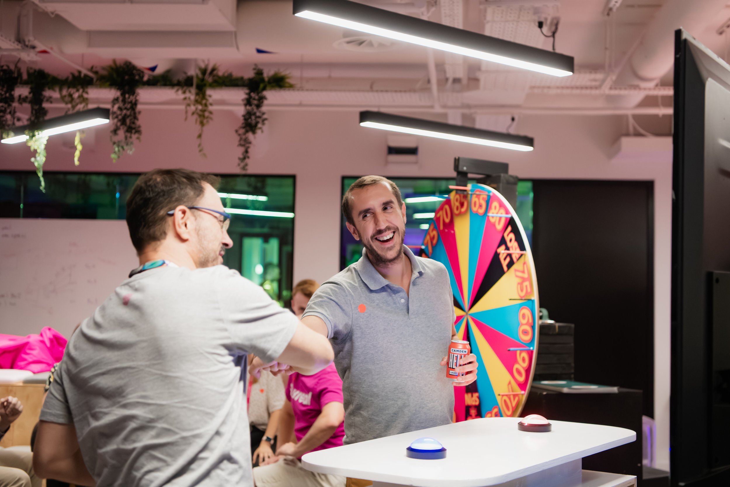 People participating in a game show with a spinning wheel behind them, in a modern indoor space with hanging plants and bright lighting.