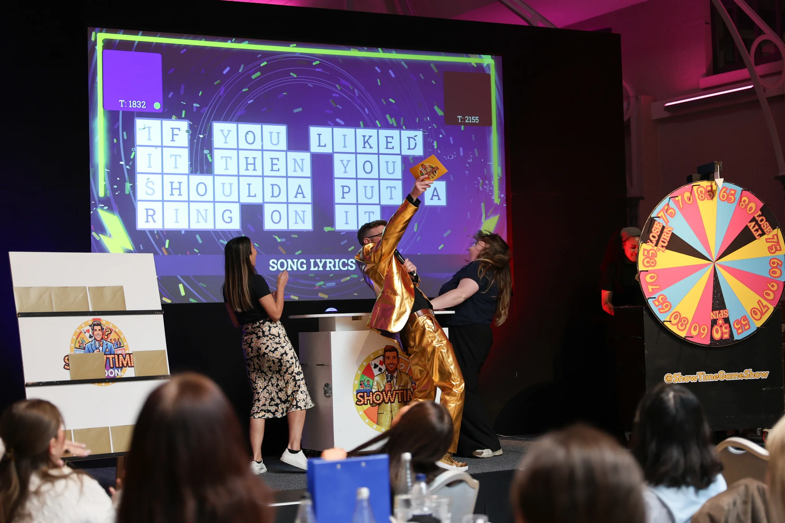 A game show setting with a large screen displaying a word puzzle, a spinning wheel on the right, and a host wearing a shiny gold jacket holding a card, engaging with a contestant. Audience members are seated in the foreground.