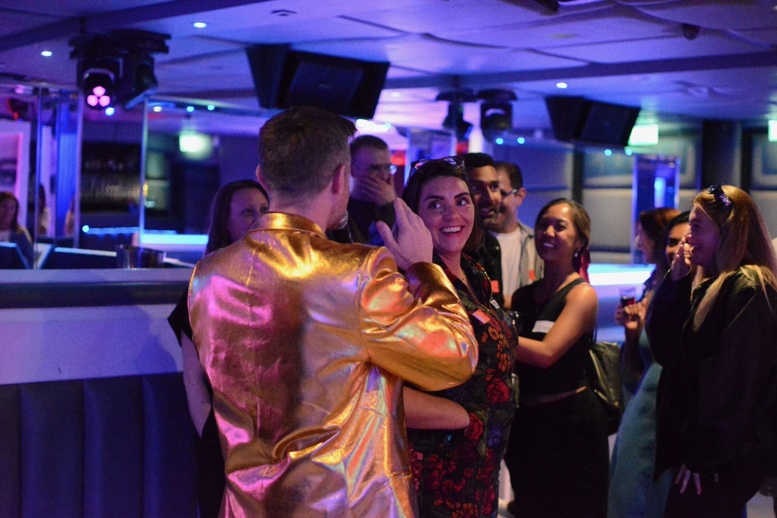 Group of people socialising at a party with colorful lighting, some wearing name tags, and a woman smiling at a man in a shiny gold jacket.