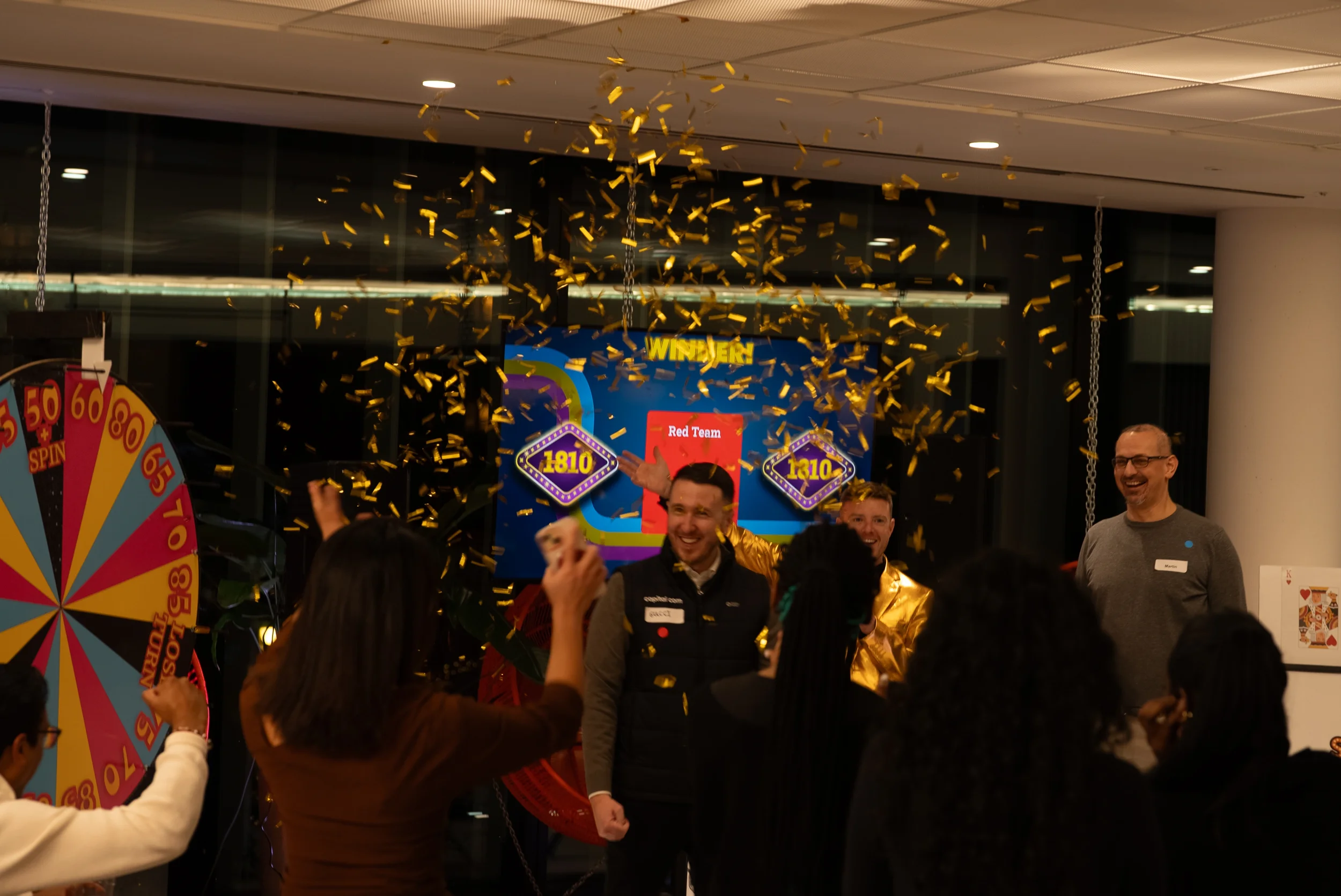 People celebrating with gold confetti in an indoor space, with a spinning wheel and prize boards visible in the background.
