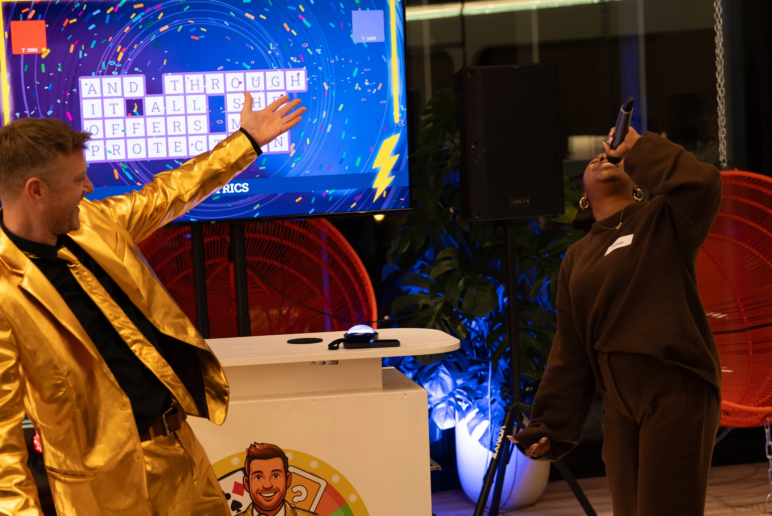 A man in a shiny gold jacket and a woman with large hoop earrings and a brown sweater enjoying a karaoke session indoors. The man points at a screen displaying a word puzzle, and the woman sings into a microphone. The background has decorative orange fans and green plants.