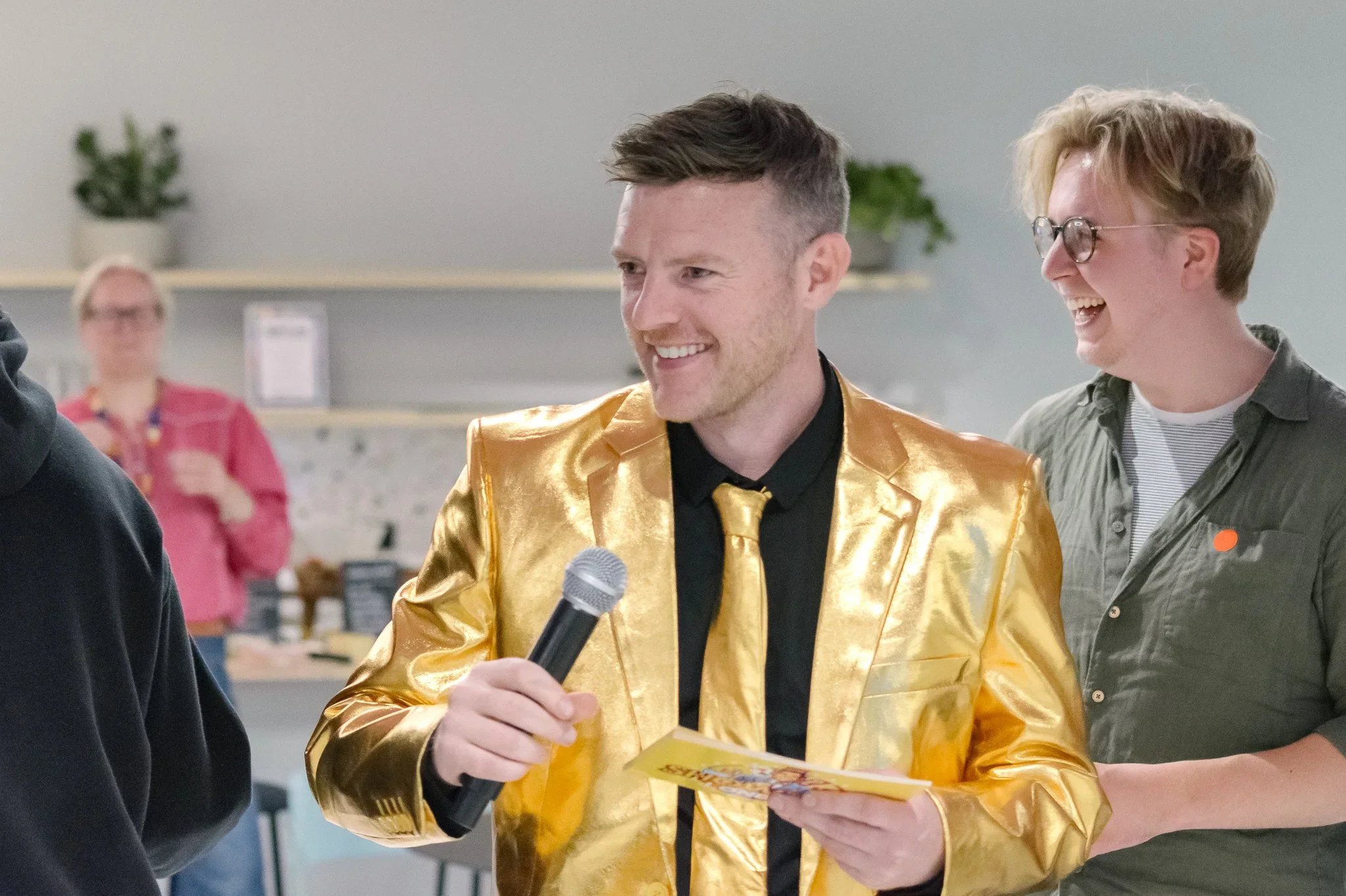 People smiling and talking at an indoor event, one person holding a microphone and a flyer, wearing a shiny gold jacket.