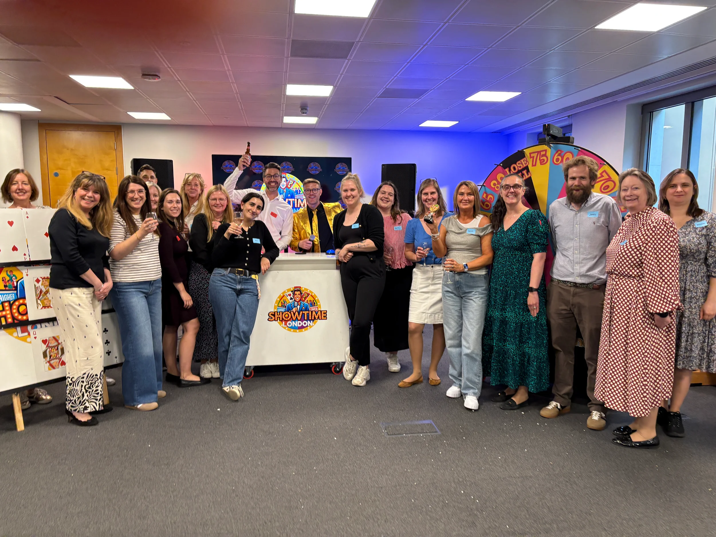 Group of people celebrating at an office party, standing in front of a Showtime London booth and a prize wheel, with some holding drinks and smiling.