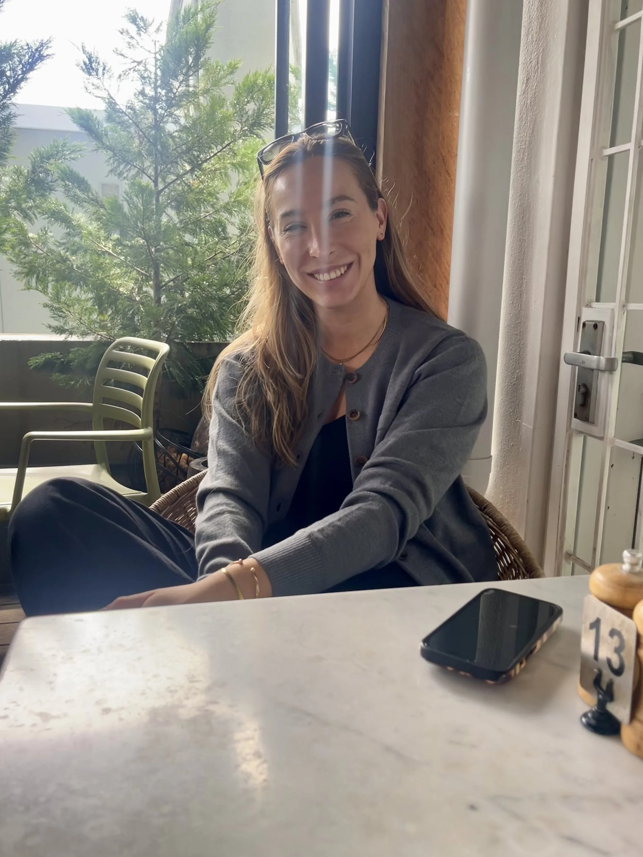 A woman with long wavy hair and sunglasses on her head, smiling and sitting at a table in a cafe with natural light and green plants in the background.
