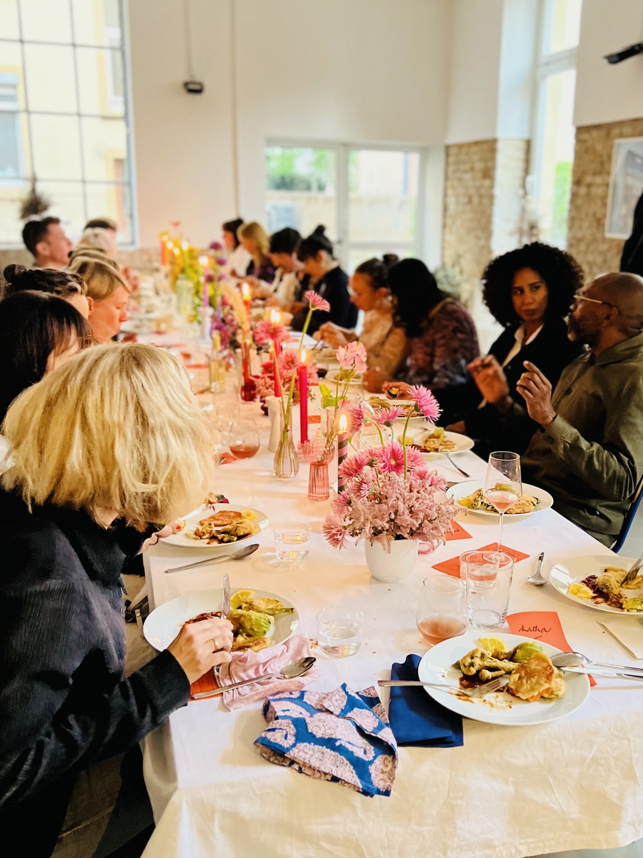 People sitting at a long dining table in Frankfurt with pink flowers, candles, and plates of food in a bright room with large windows.