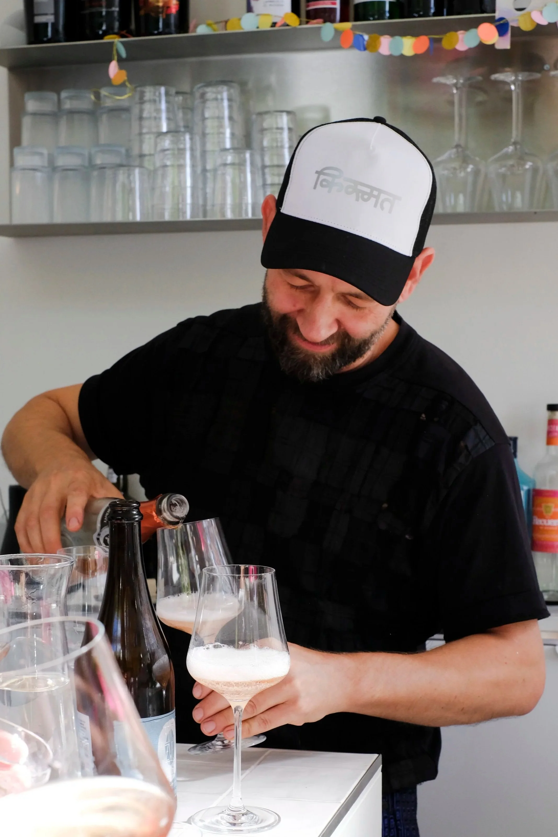 A man with a beard and dark hair wearing a black shirt and a black and white cap pouring a drink into a wine glass at a bar or gathering, with several glasses and bottles on the table and shelves with glasses and bottles in the background.