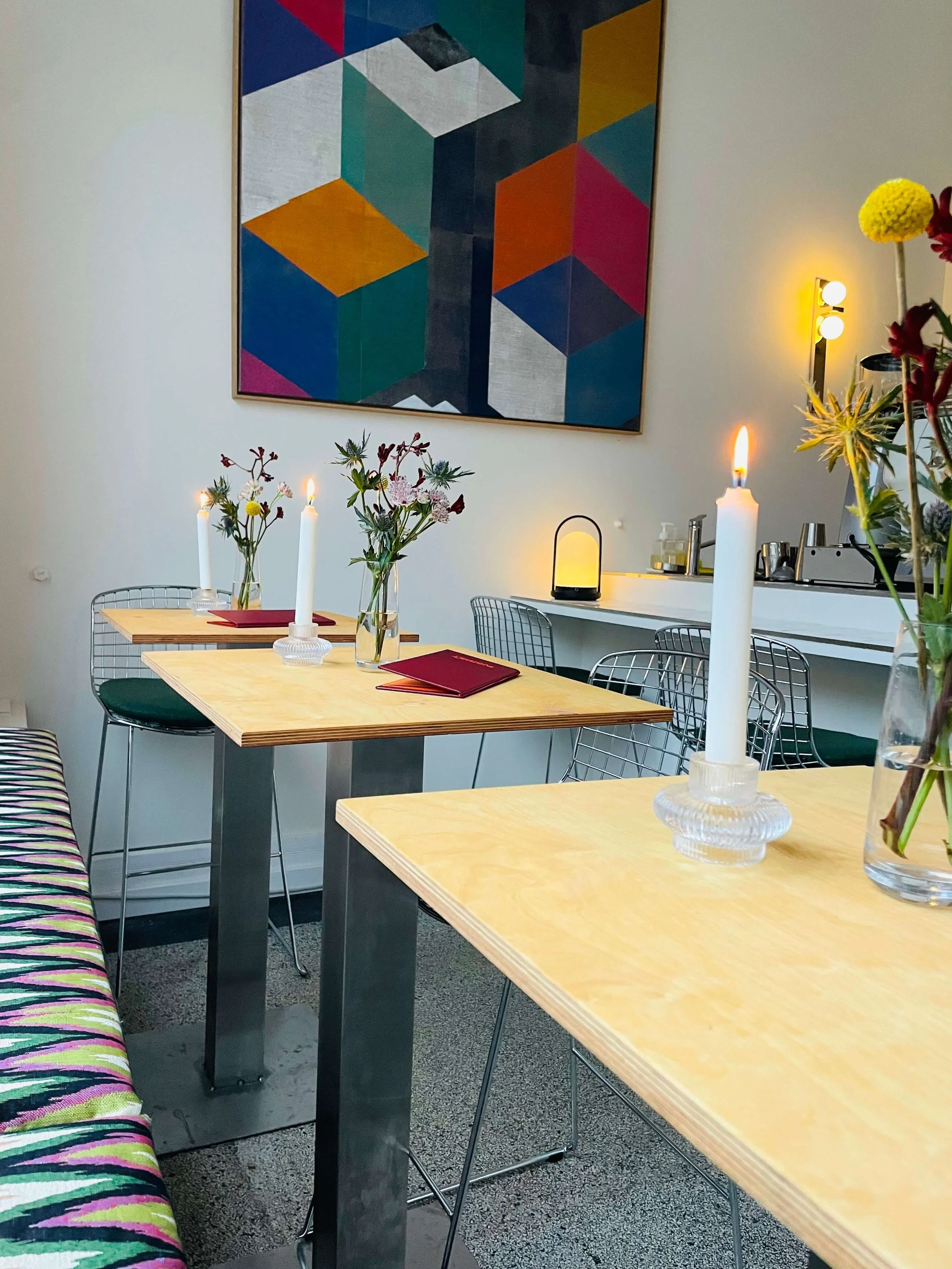 A cozy dining area with wooden tables decorated with candles and flowers, modern metal chairs, and a colorful geometric abstract painting on the wall.