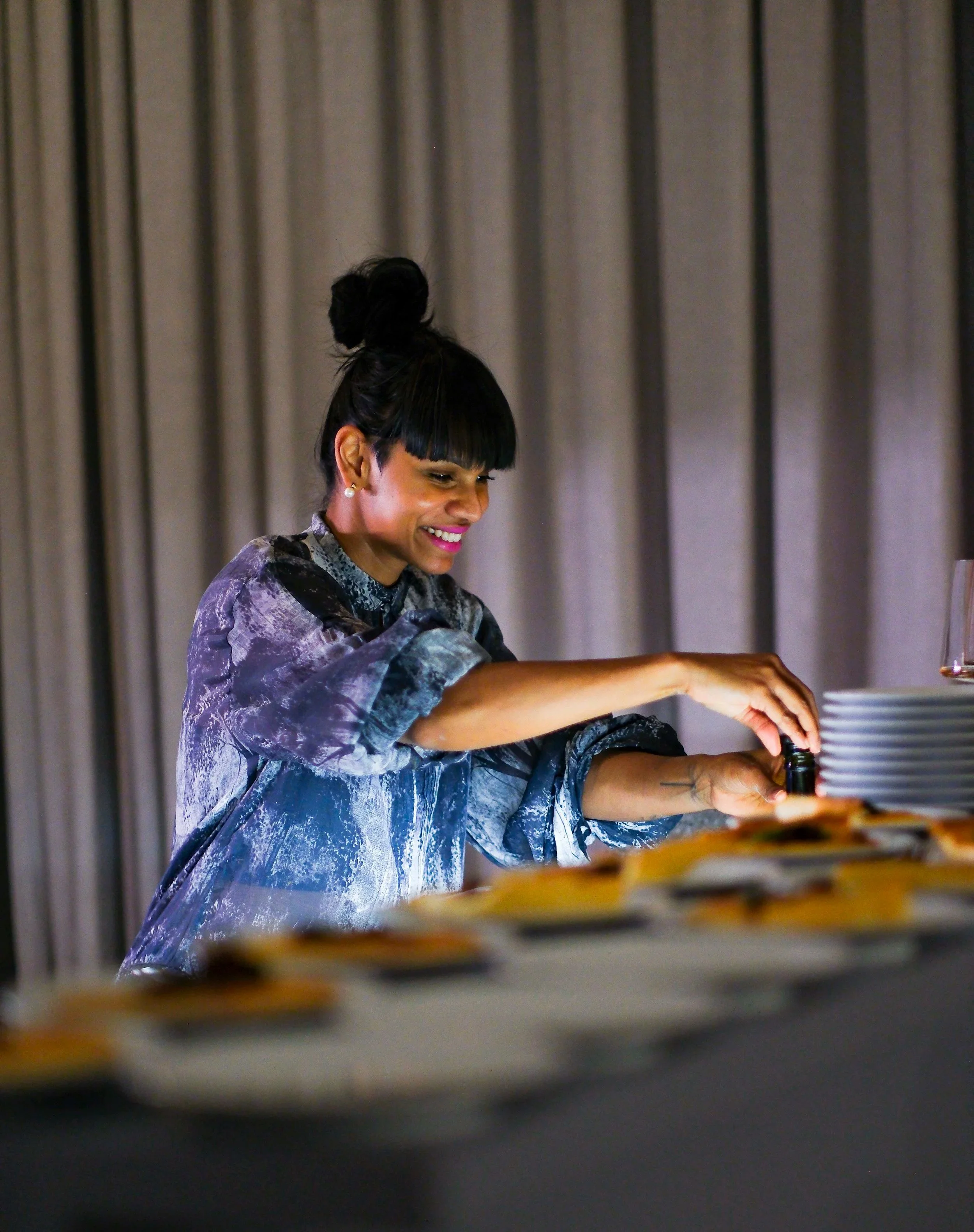 A woman with dark hair in a bun, wearing a patterned blouse, smiling as she prepares food at a buffet table with stacks of plates and glasses.