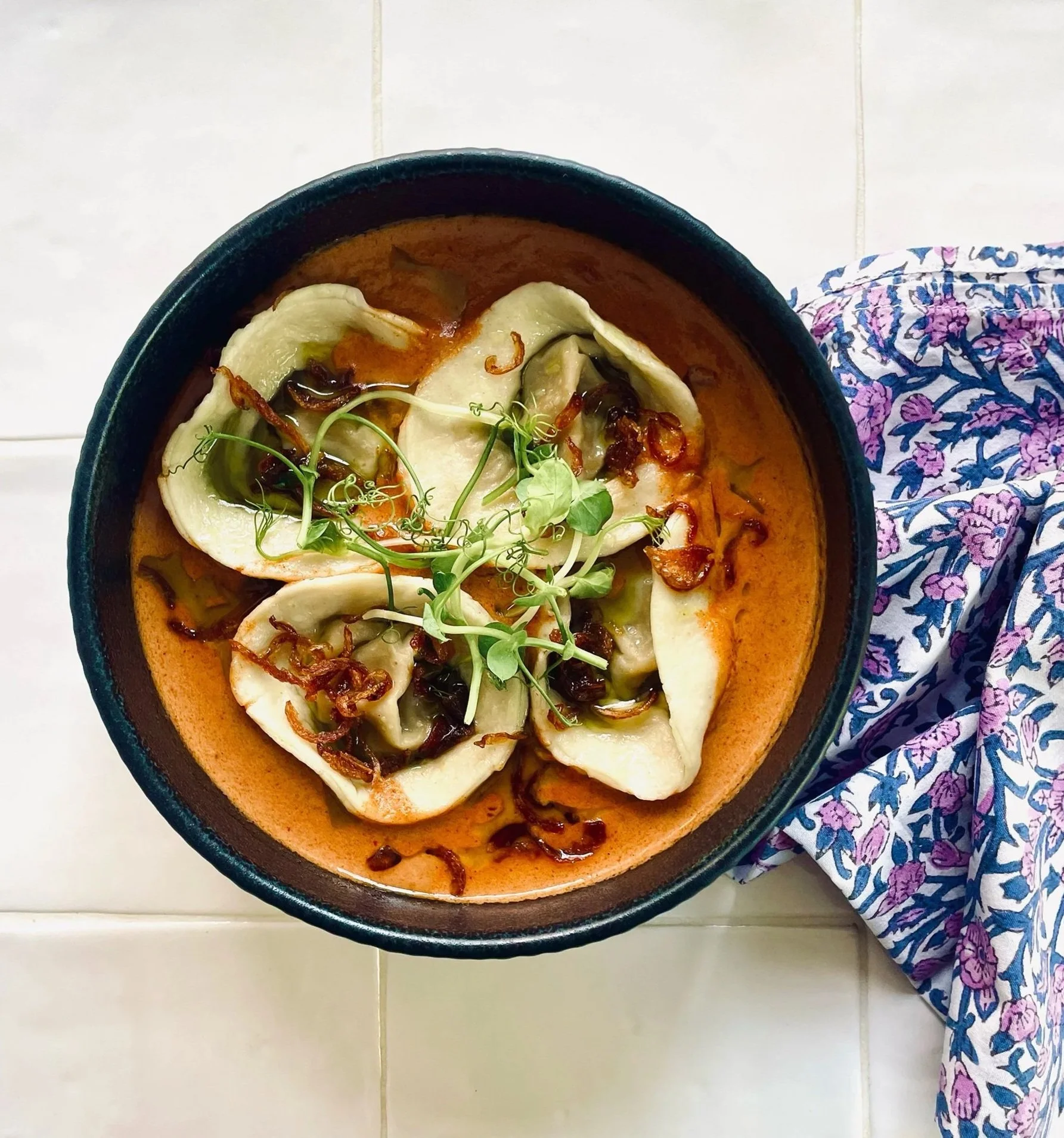 Bowl of hand-made dumplings with sauce, garnished with fried shallots and microgreens on a white tiled surface with a blue floral cloth nearby.