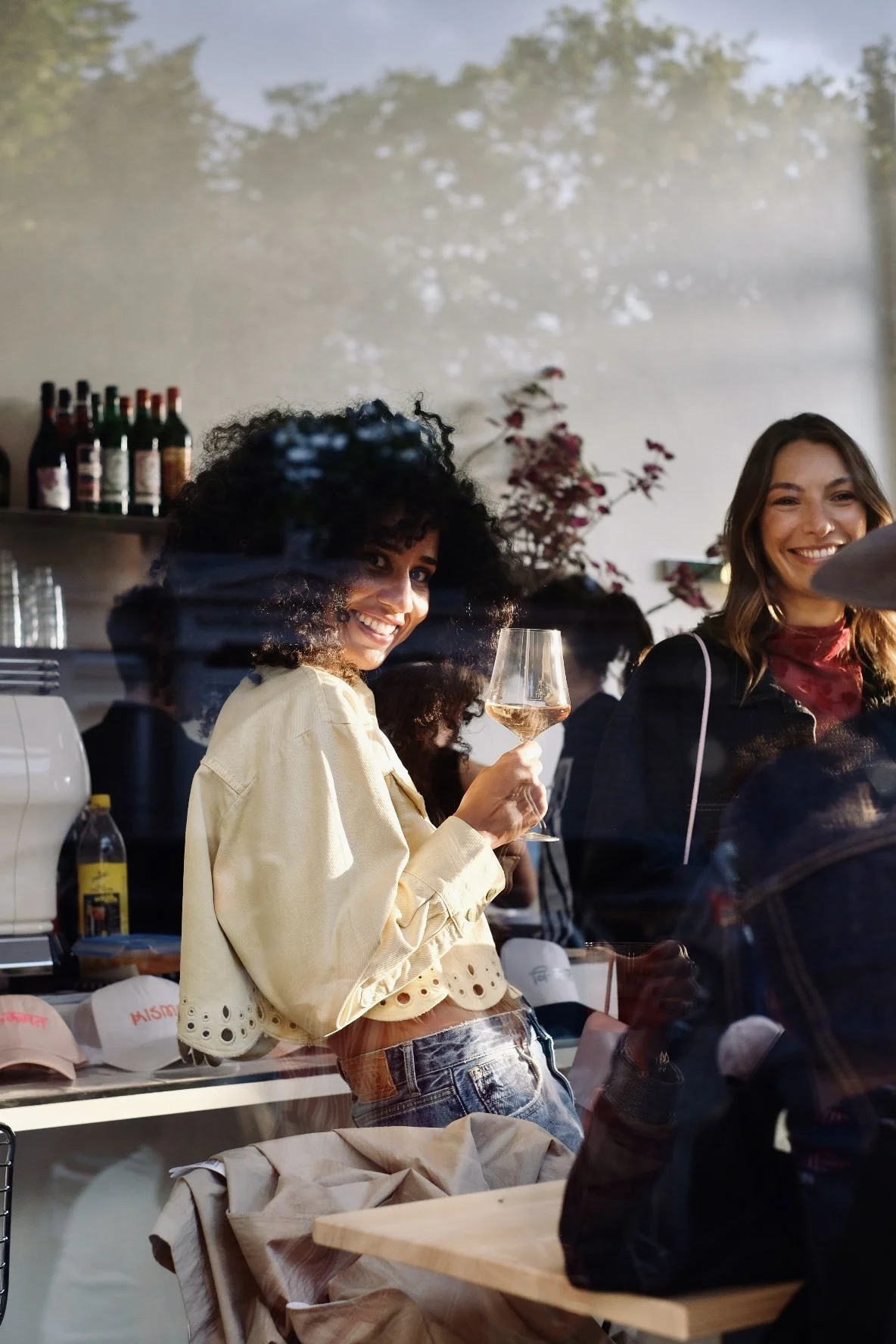 Two women smiling, one holding a glass of white wine, inside a cozy cafe or restaurant with a glass window reflecting trees outside.