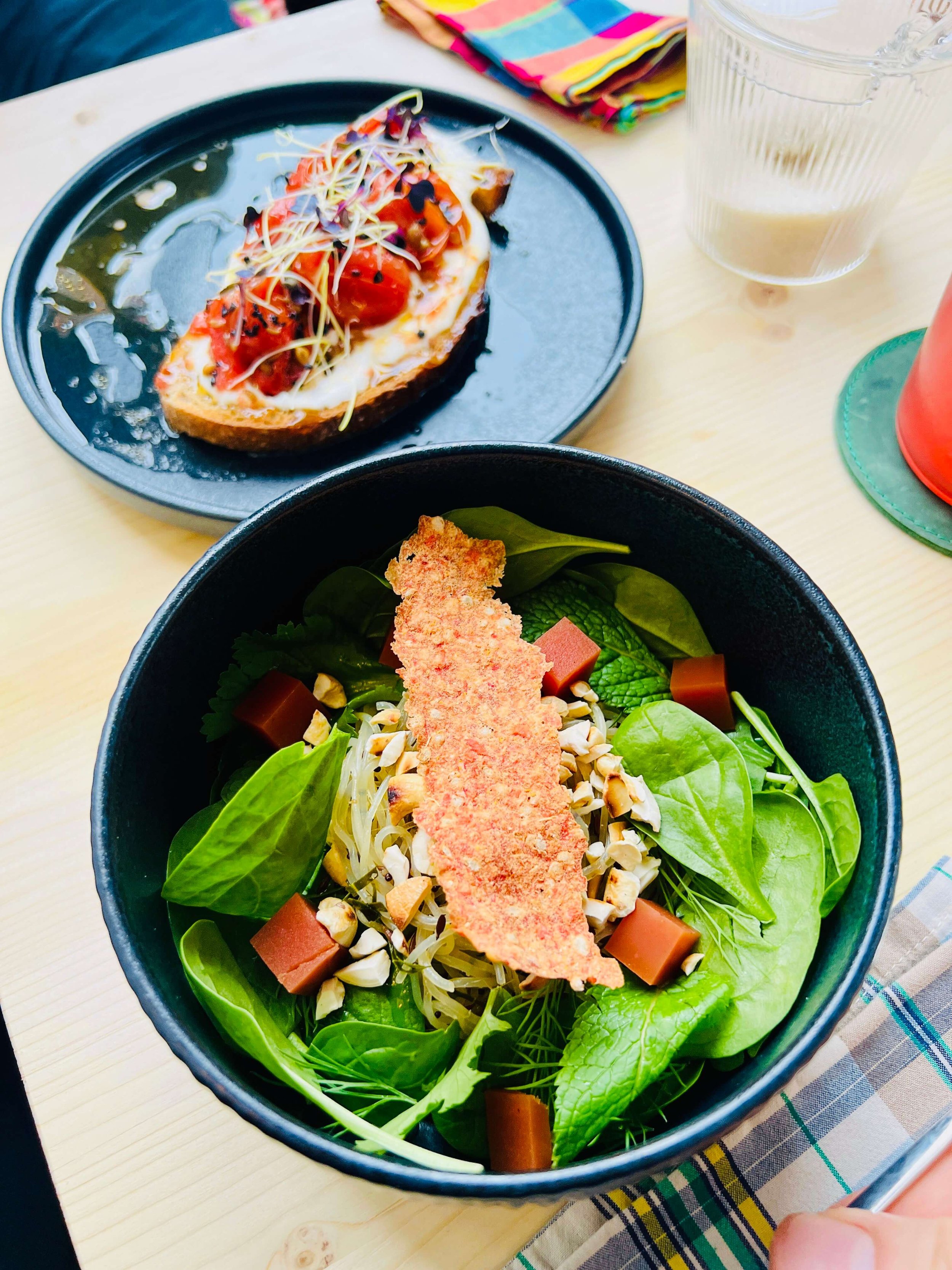 A salad in a black Playground bowl with mixed greens, chopped nuts, cubed cheese, and a crispy breadstick on top, next to a toast topped with cherry tomatoes and whipped feta, and sprouts, with drinks and a napkin nearby.