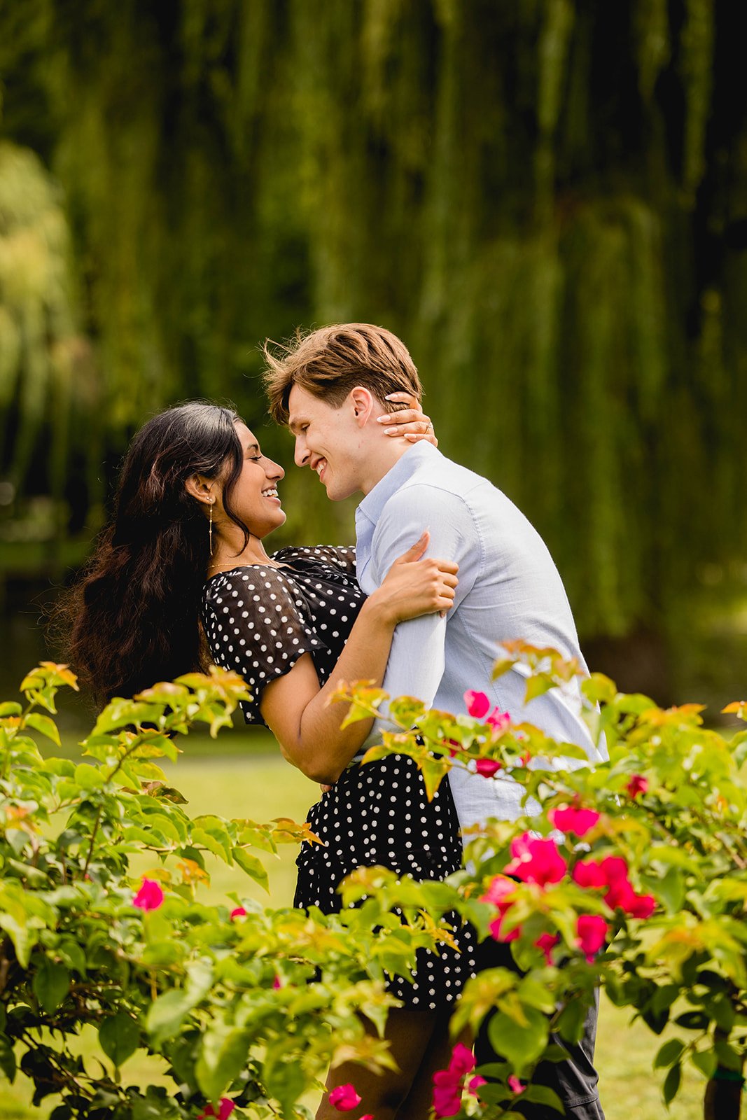 Couple smiling and embracing among greenery and willow trees during a Boston Public Garden engagement photo session.