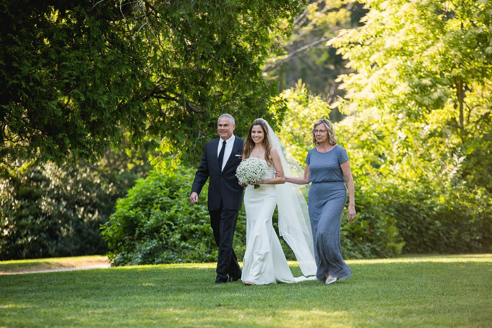 Bride walking down the aisle with her parents during outdoor wedding ceremony at Estate at Moraine Farm in Beverly, Massachusetts