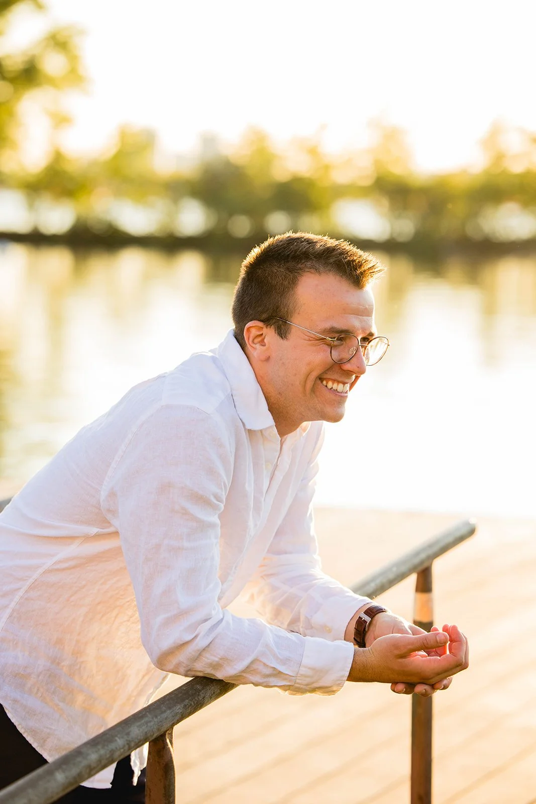 A man with short dark hair, glasses, and a white shirt leaning on a railing near a body of water, smiling during sunset.