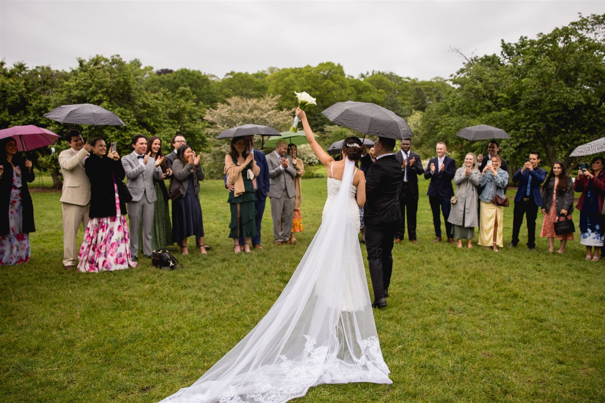 newly wed couple standing under umbrella facing their wedding guests celebrating their accomplishment at Arnold's Arboretum in Jamaica Plains Boston