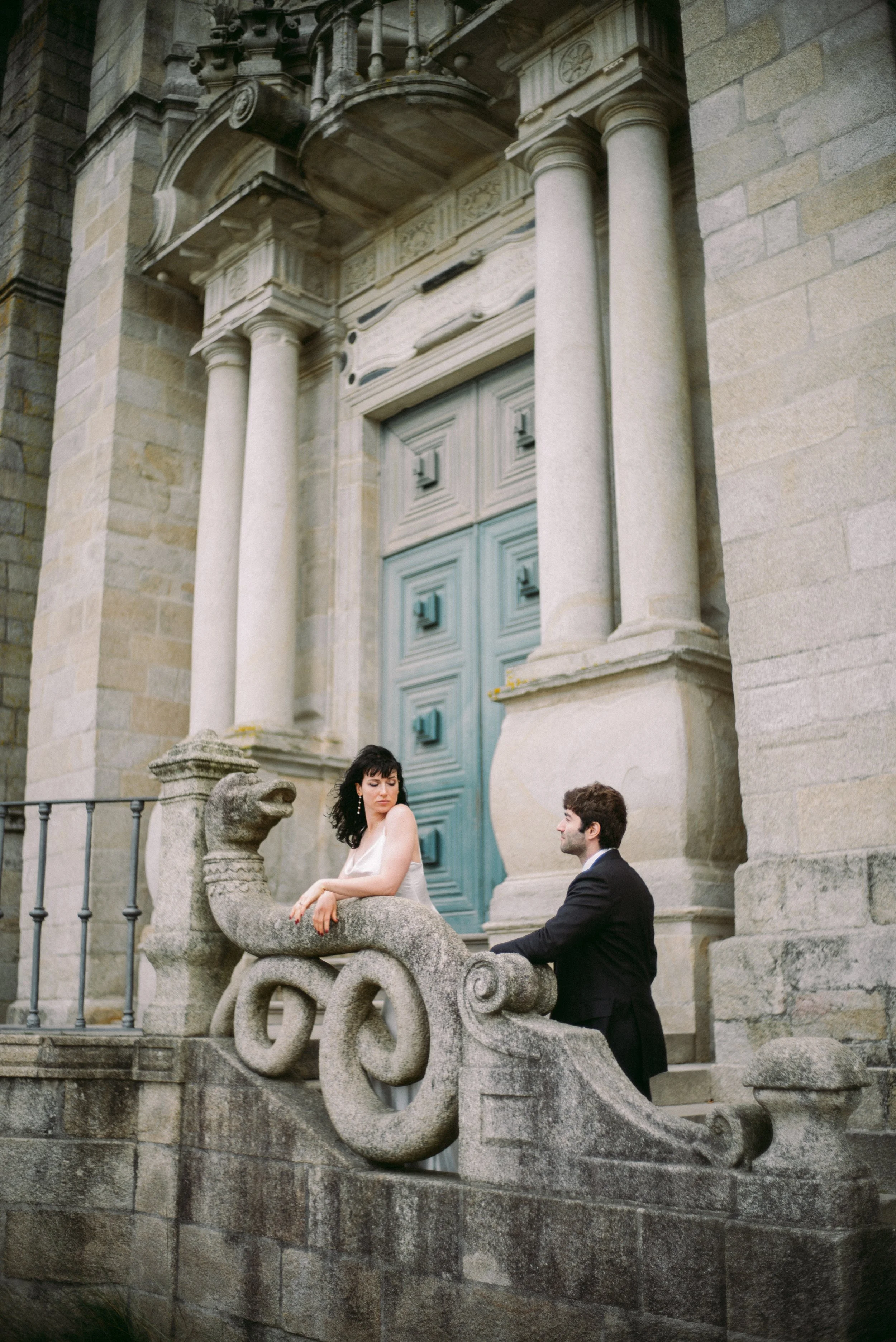 A man in a suit kneeling in front of a woman in a white dress sitting on a stone sculpture of a dragon on an ornate stone staircase outside a historic building with large wooden doors and architectural details.