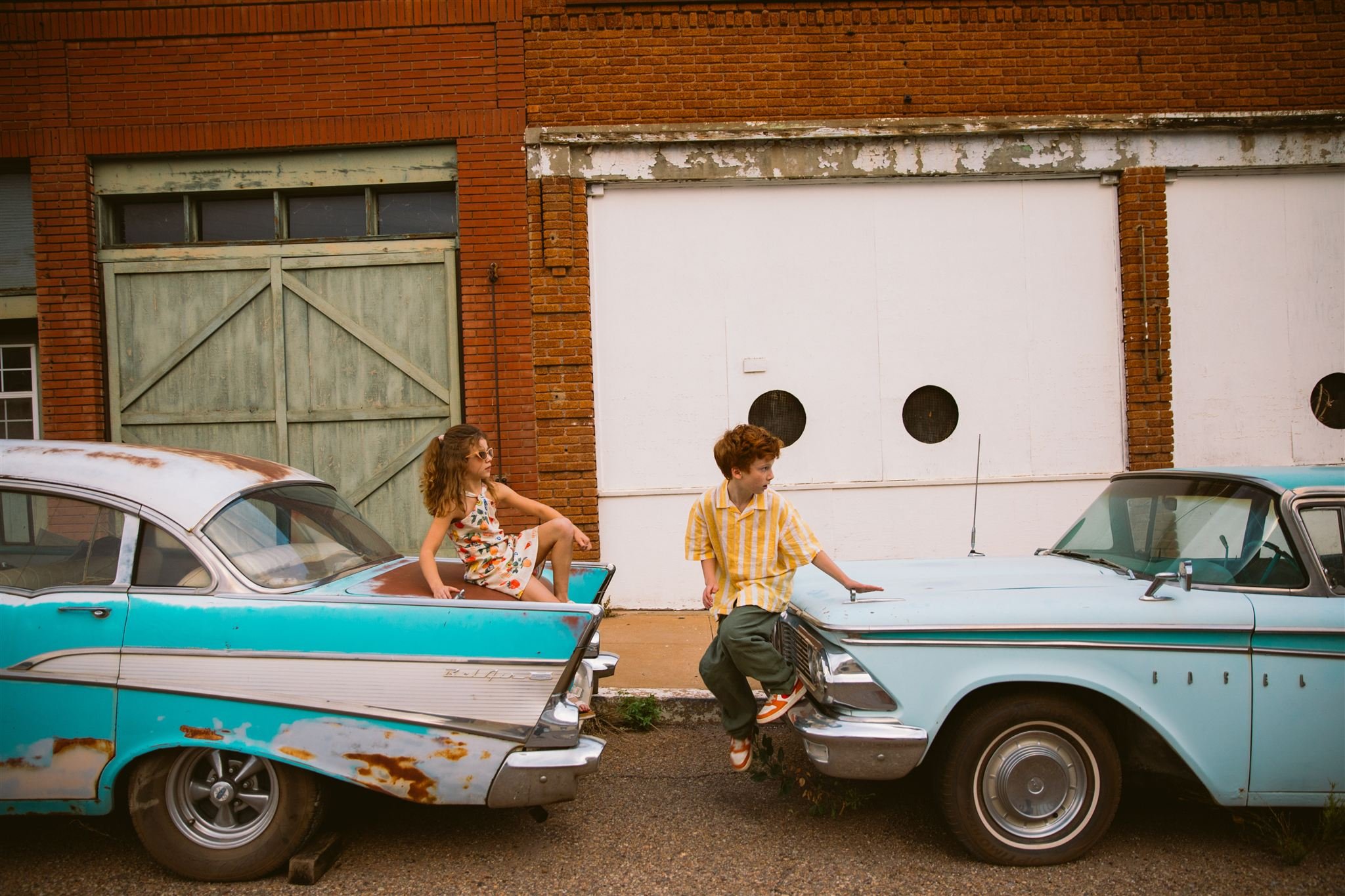 Cinematic family photography in Bisbee, Arizona, showing children sitting between two vintage cars on a quiet historic street with colorful brick buildings