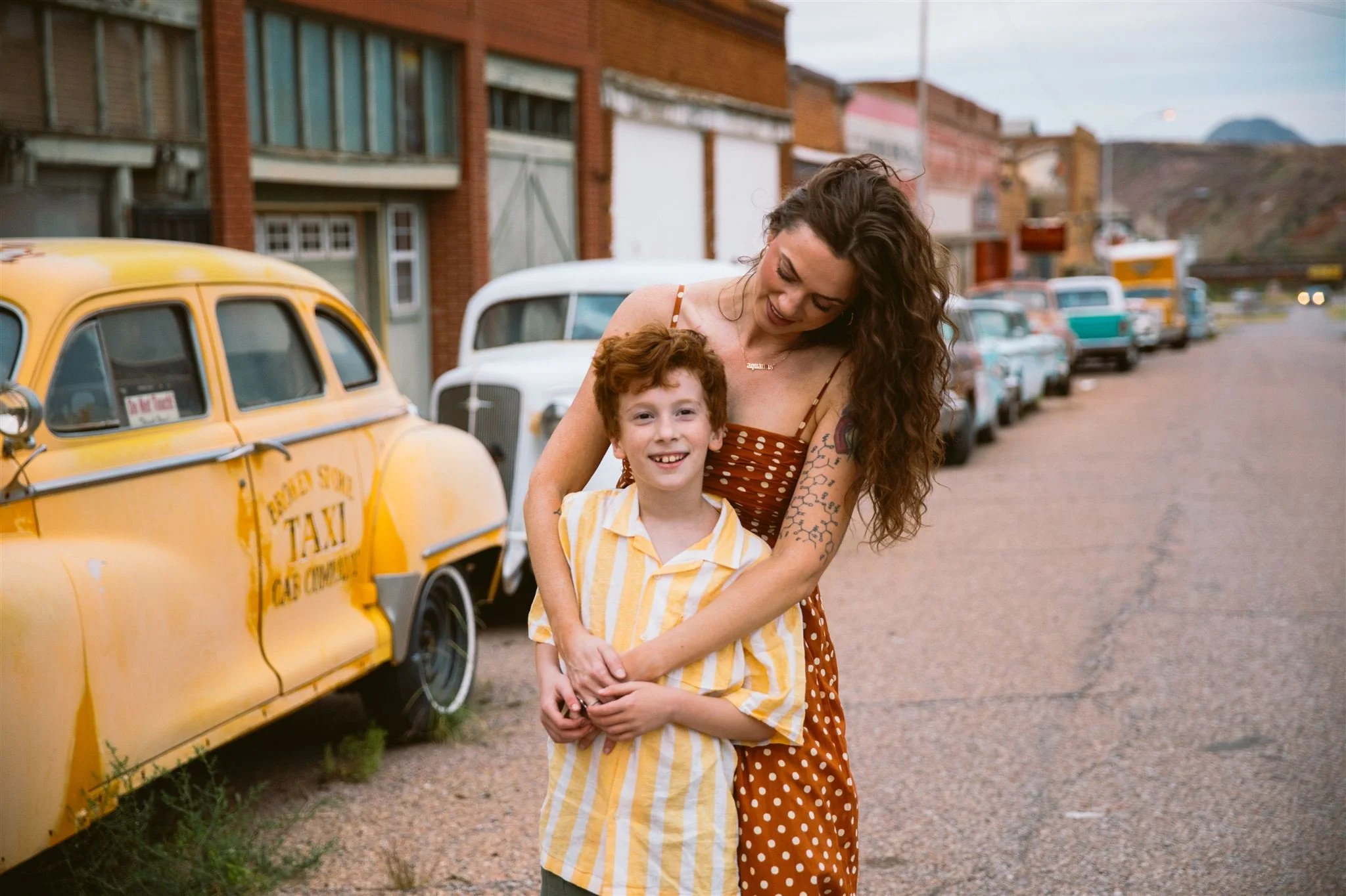 Cinematic family photography session in Bisbee, Arizona, showing a mother and son smiling together on a quiet historic street with vintage cars and small-town storefronts