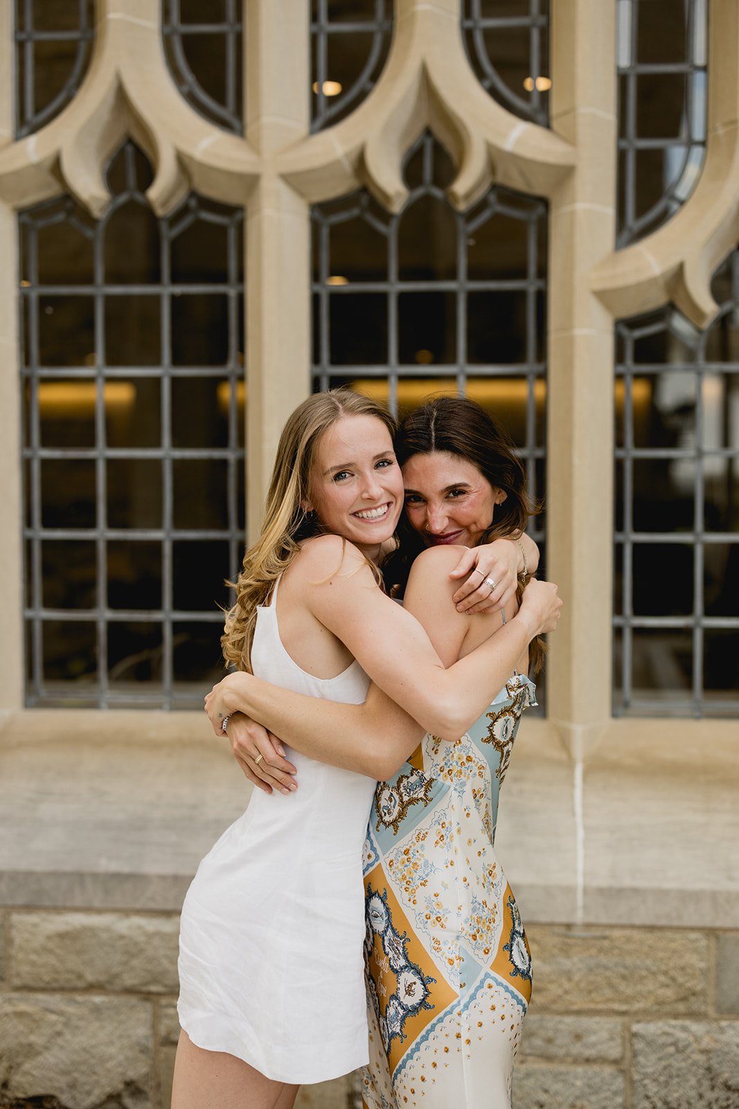 Boston College graduates hugging in front of a beautiful window on the school campus