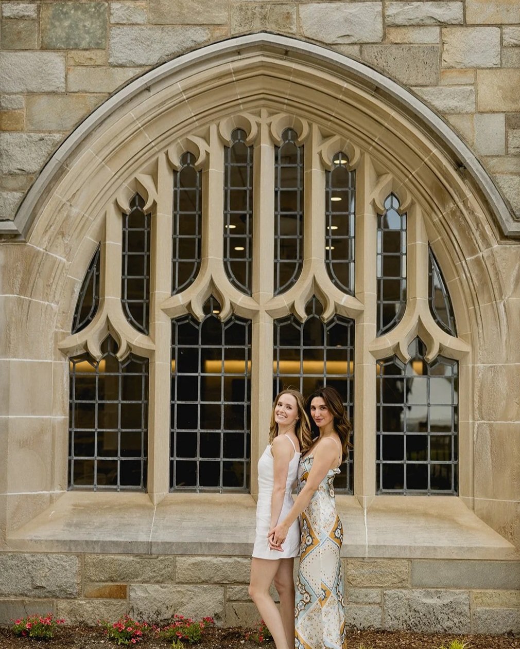 Boston College graduates posing and smiling in front of a beautiful window on campus
