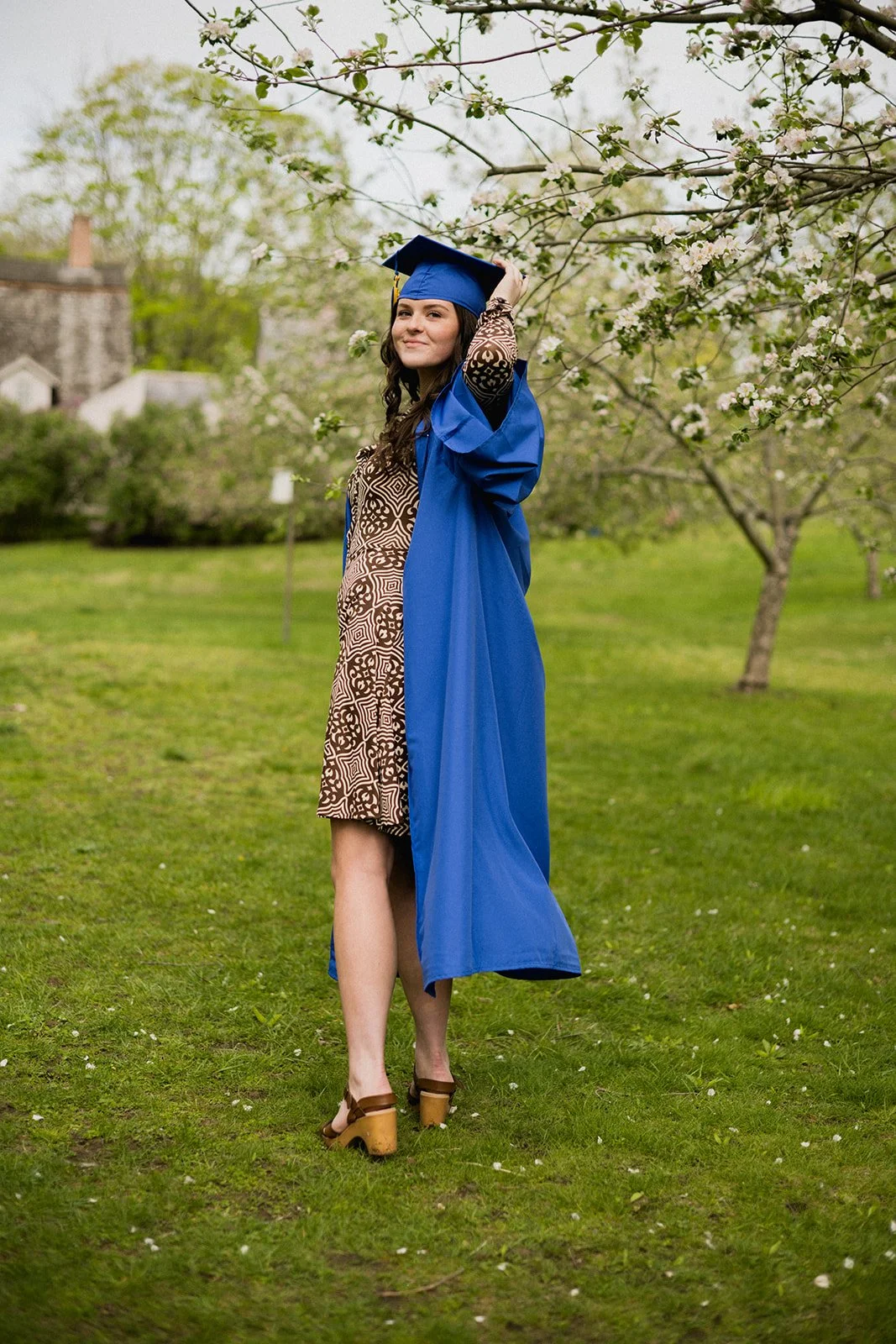 High school senior in a blue cap and gown standing among flowering trees during a spring senior photo session in Massachusetts.