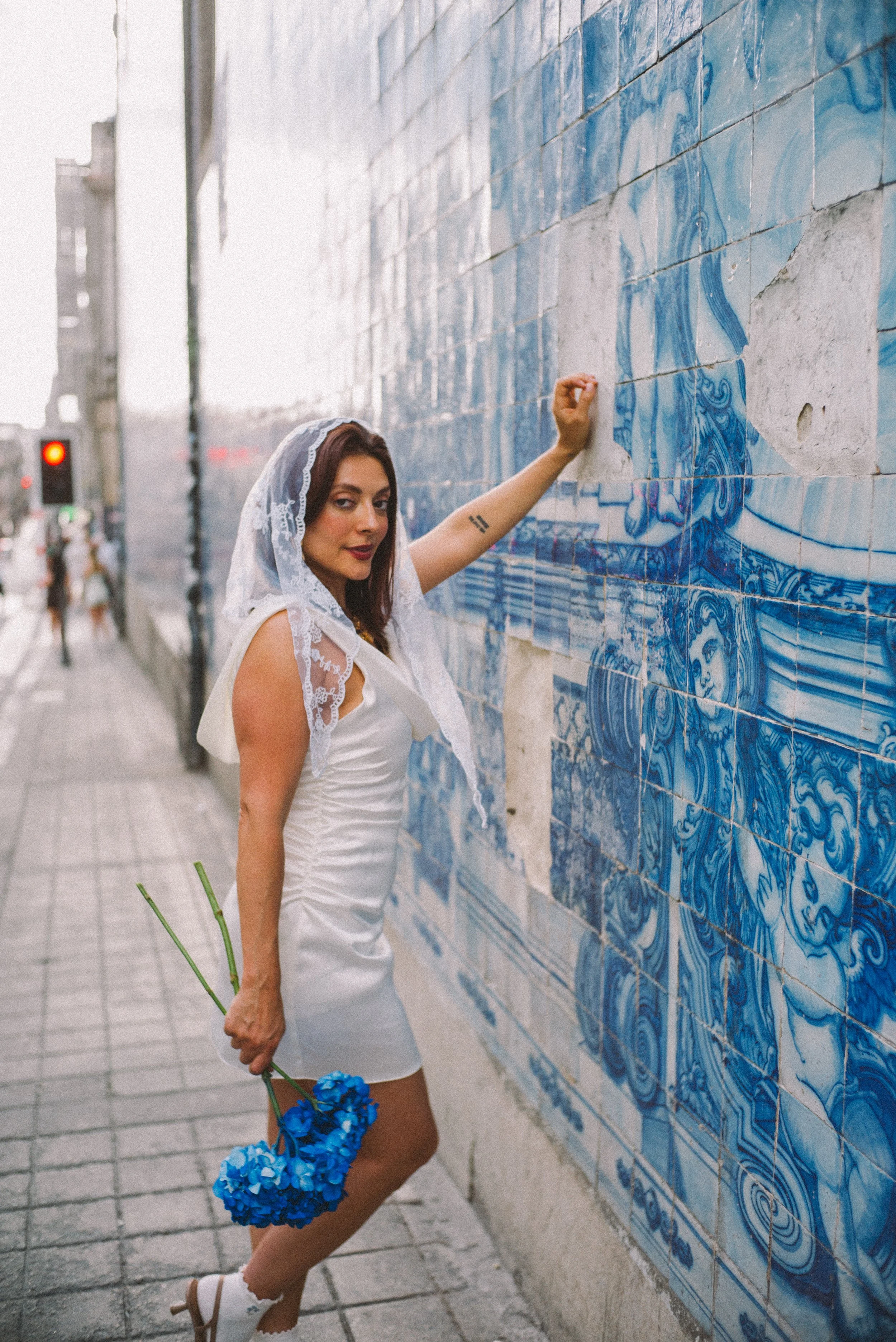 A woman dressed in white with a lace head covering stands against a wall of blue and white patterned tiles on a city street. She holds blue flowers in one hand and gently touches the wall with her other hand, gazing at the camera.
