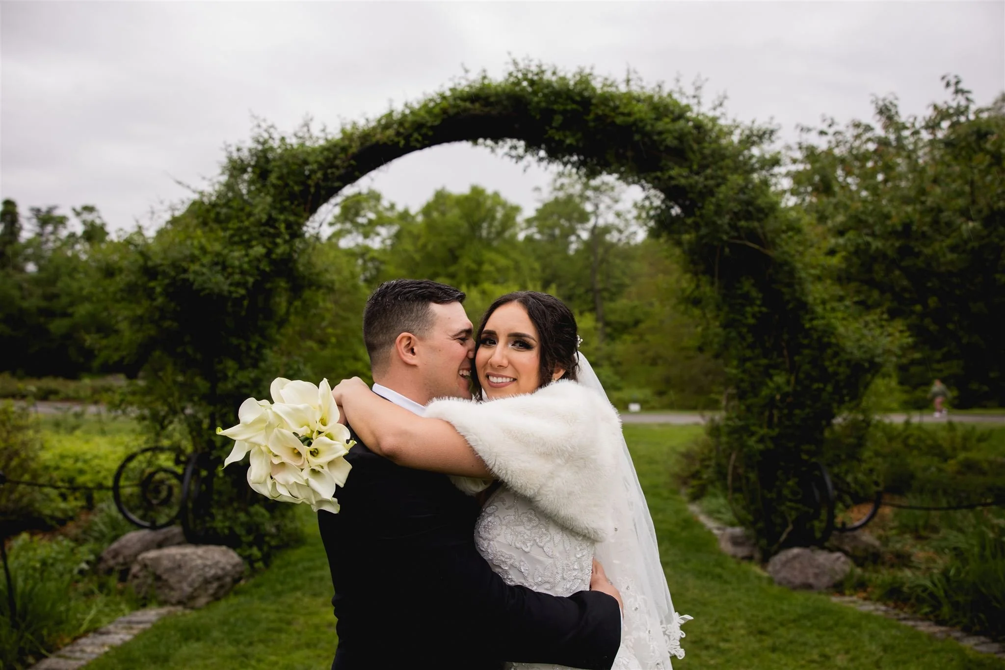 newly wed couple smiling candidly and embracing underneath a vine-lined archway at  Arnold's Arboretum in Jamaica Plains Boston
