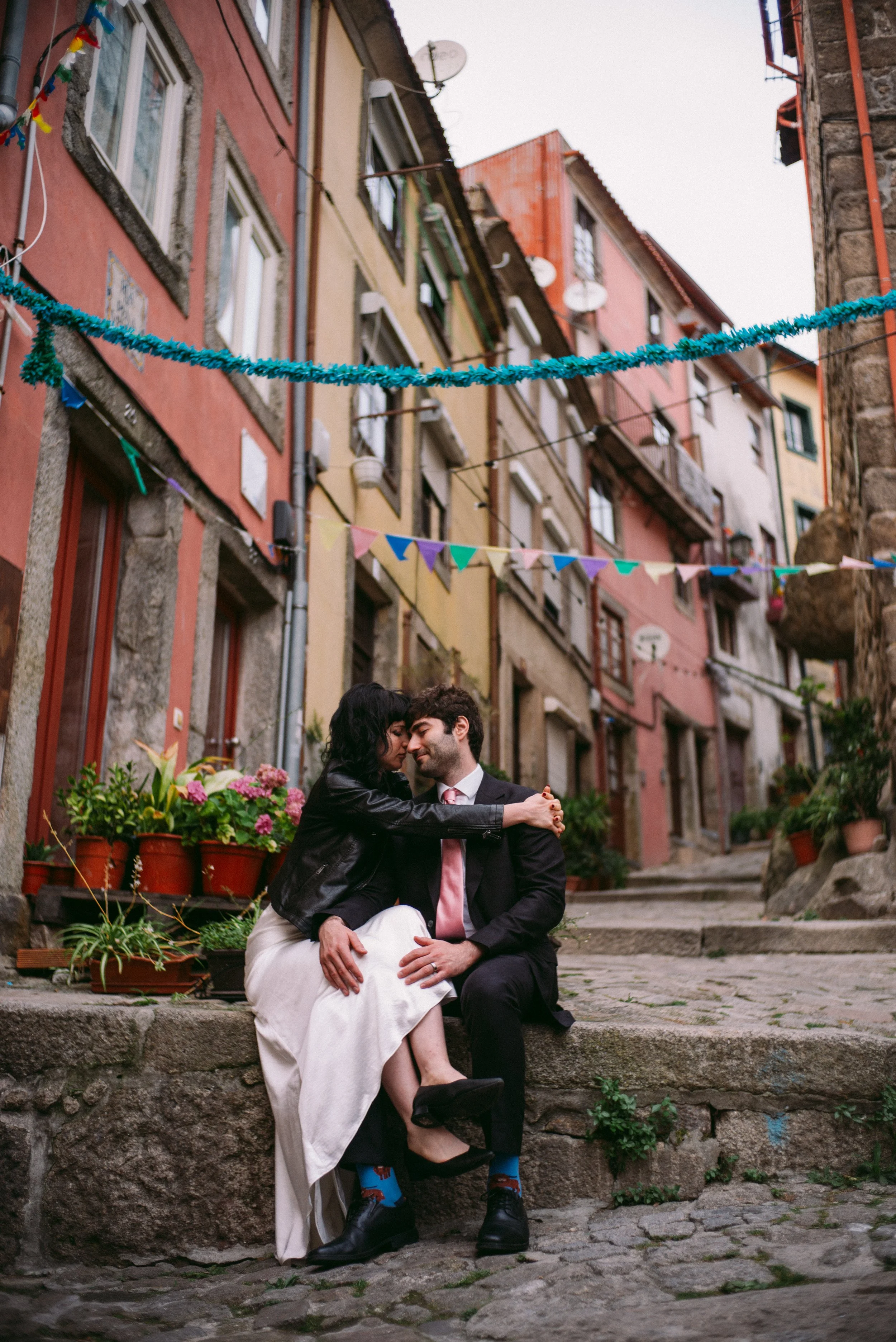 A couple sitting on stone steps in a colorful outdoor setting, holding hands and sharing an intimate moment. The background features multi-colored buildings with balconies, satellite dishes, and decorative flags hanging across the street.