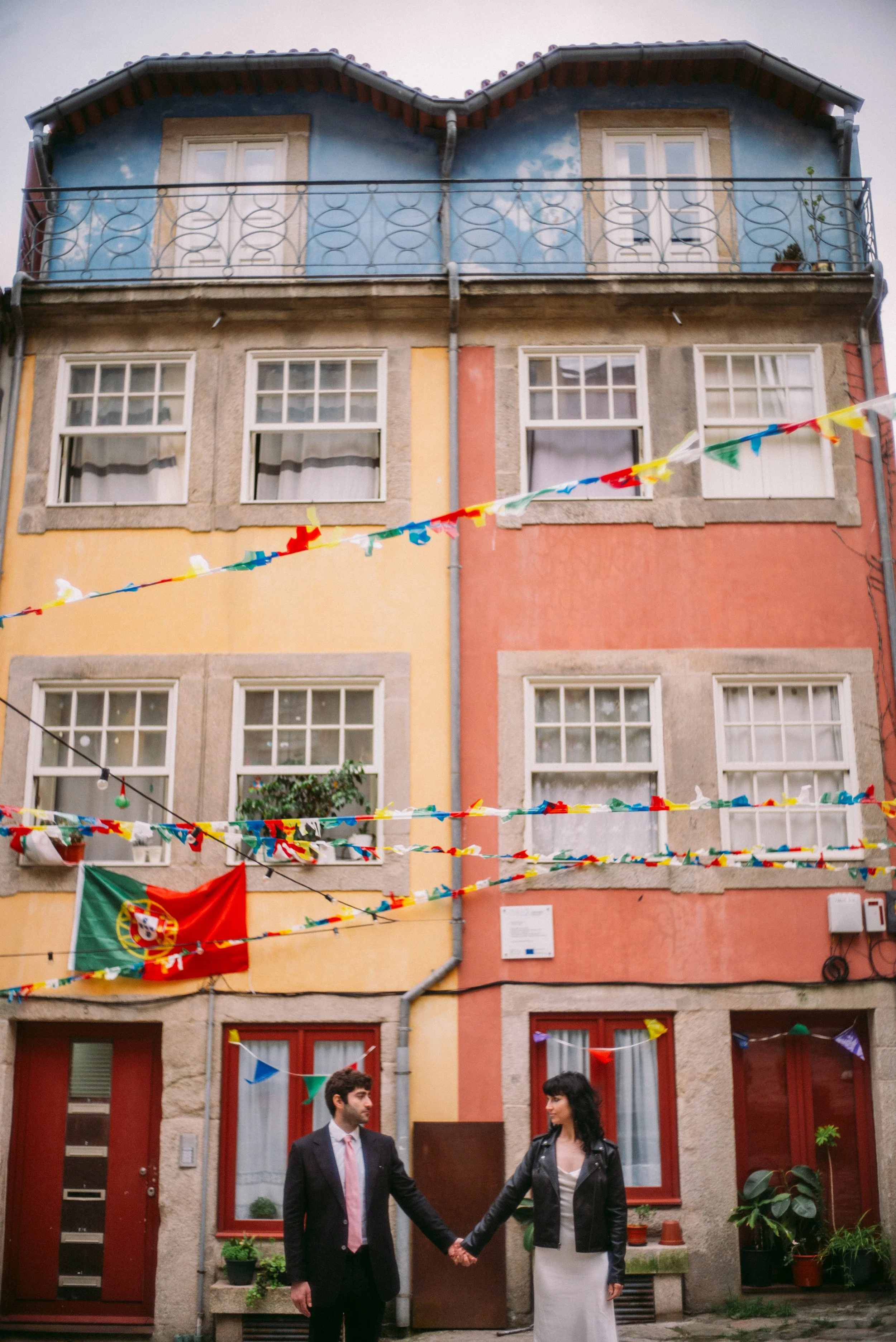A man and a woman holding hands in front of a colorful building decorated with string lights and a Portuguese flag, possibly at a celebration or event.