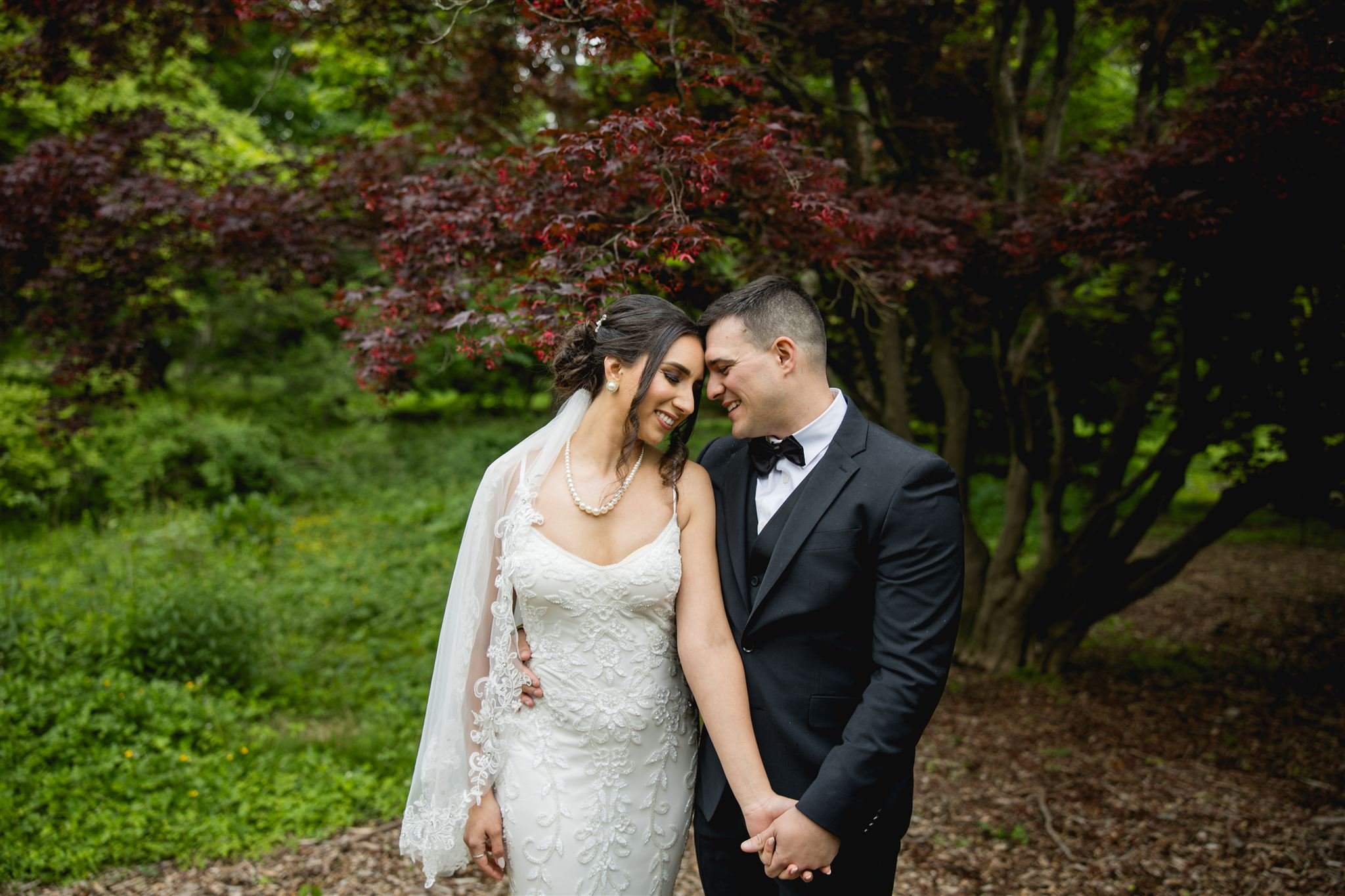 newly wed couple forehead to forehead smiling candidly with eyes closed and holding hands under a tree in a garden