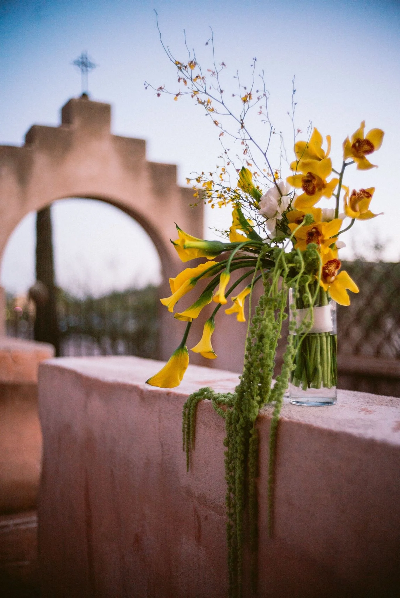 A glass vase filled with yellow and white flowers, along with green foliage, sitting on a pinkish surface with a blurred background of an arch and a cross at sunset.