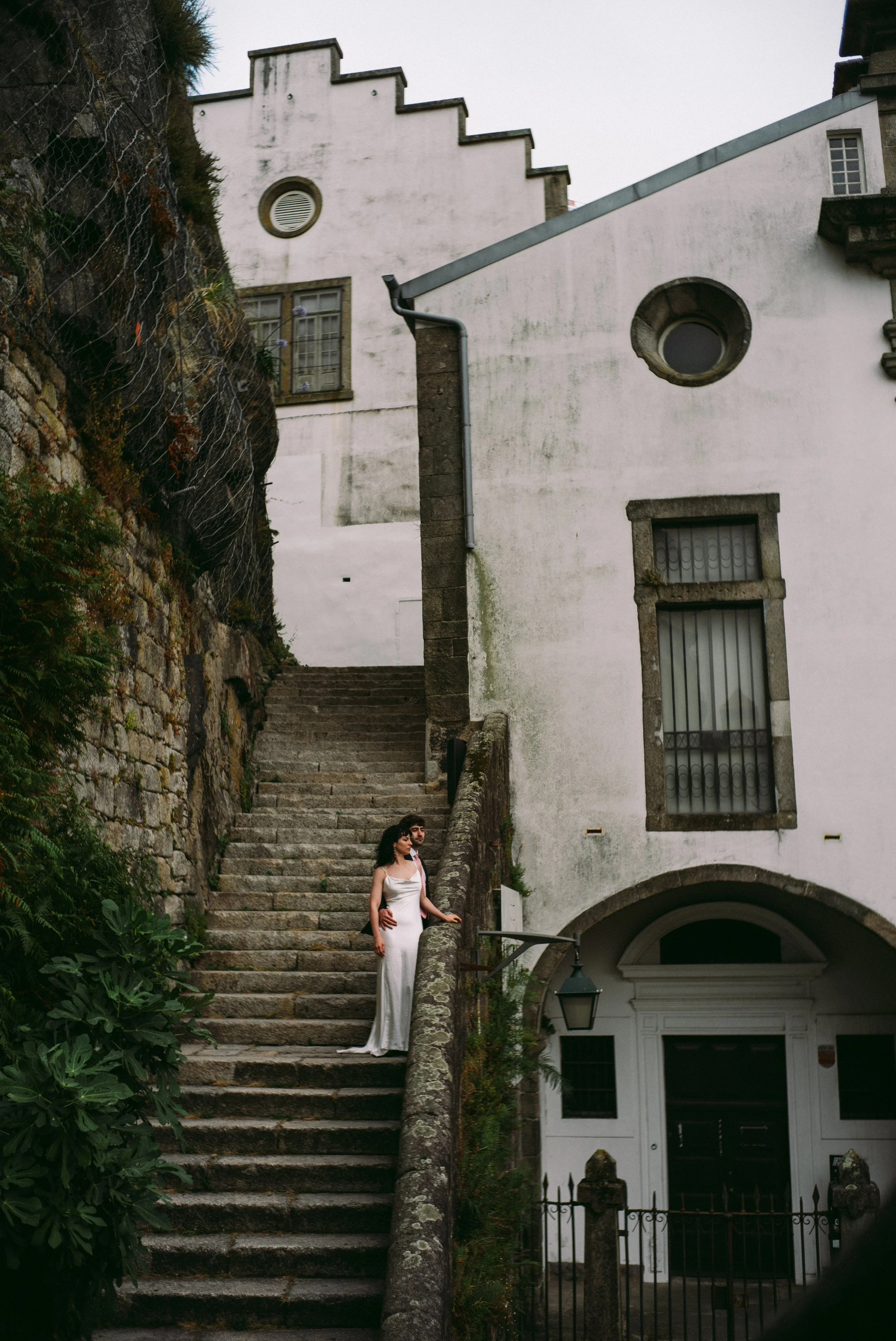 A woman in a white dress and a man in a dark jacket stand on the stone staircase outside a white building with rounded and rectangular windows, surrounded by greenery and a black wrought iron fence.