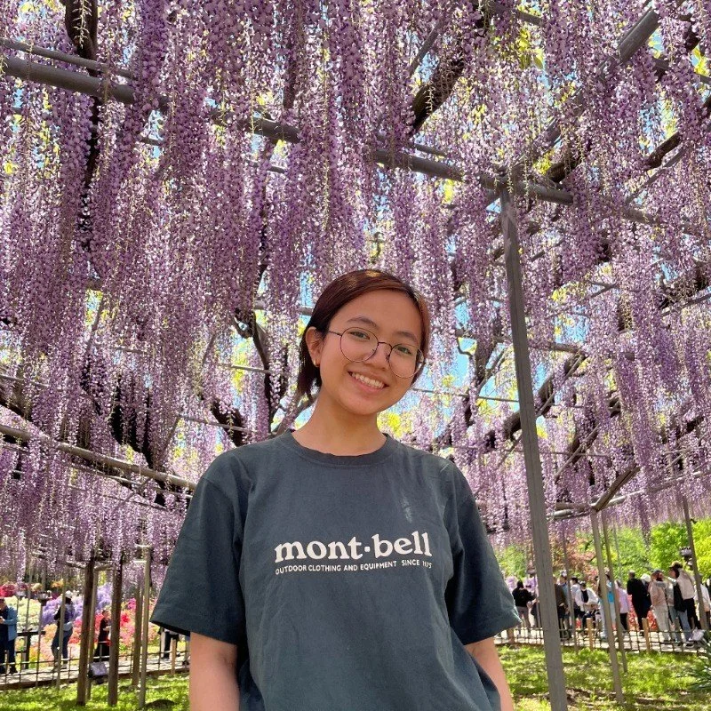 A smiling young woman with glasses and a mont-bell t-shirt standing under hanging purple wisteria flowers in a garden.