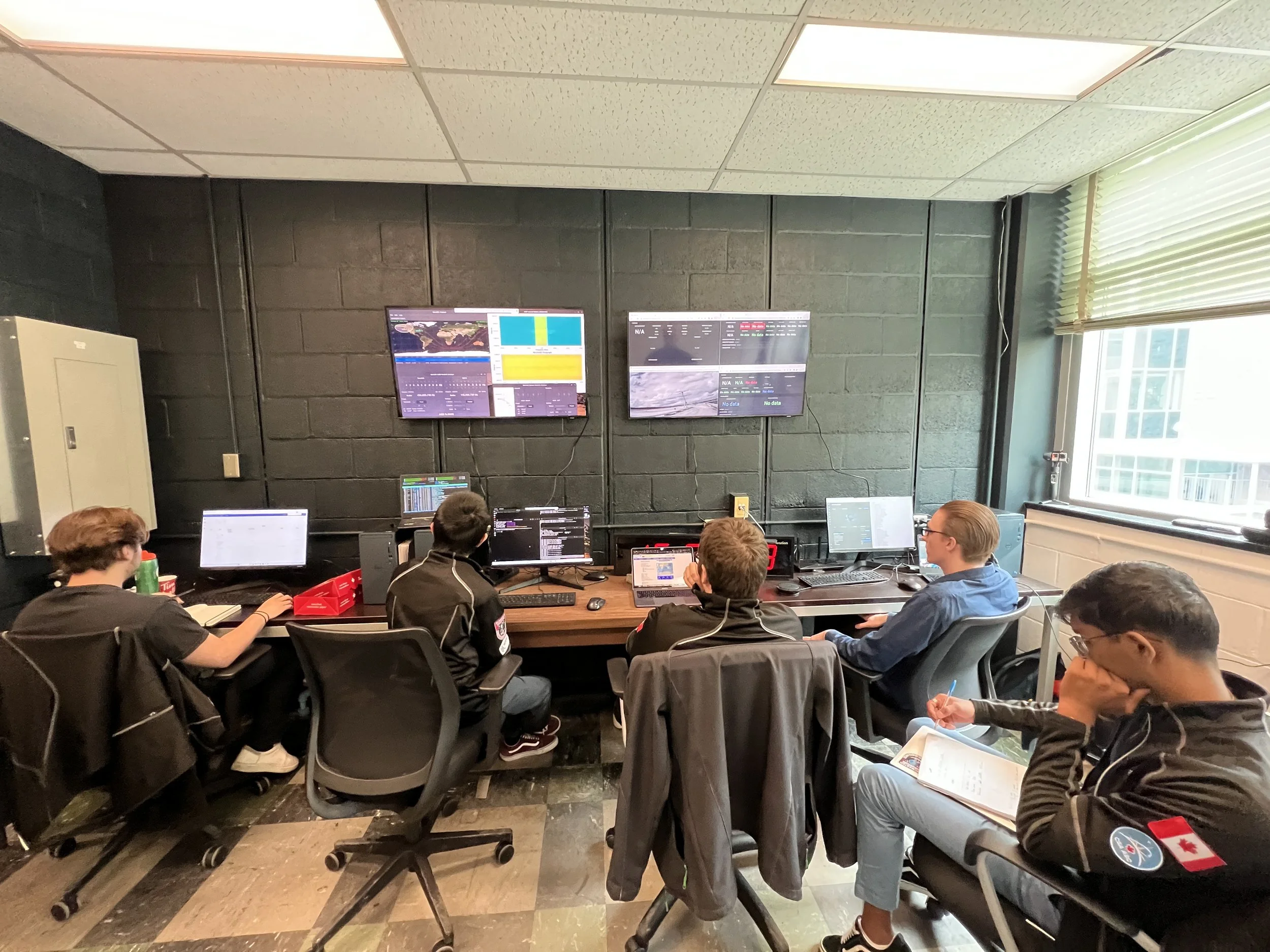 Group of five young people in a control room with multiple monitors on the wall, working at computers and taking notes, with a window on the right side letting in natural light.