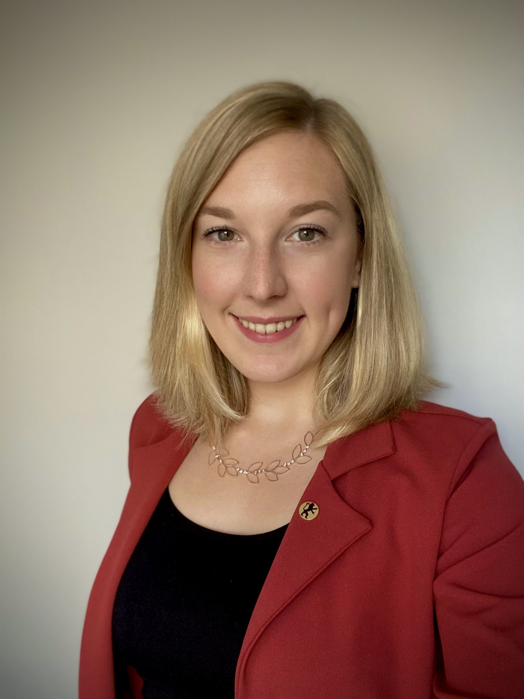 A portrait of a young woman with shoulder-length blonde hair, wearing a red blazer, black top, a silver leaf-shaped necklace, and a circular pin on her blazer, smiling against a plain light-colored background.