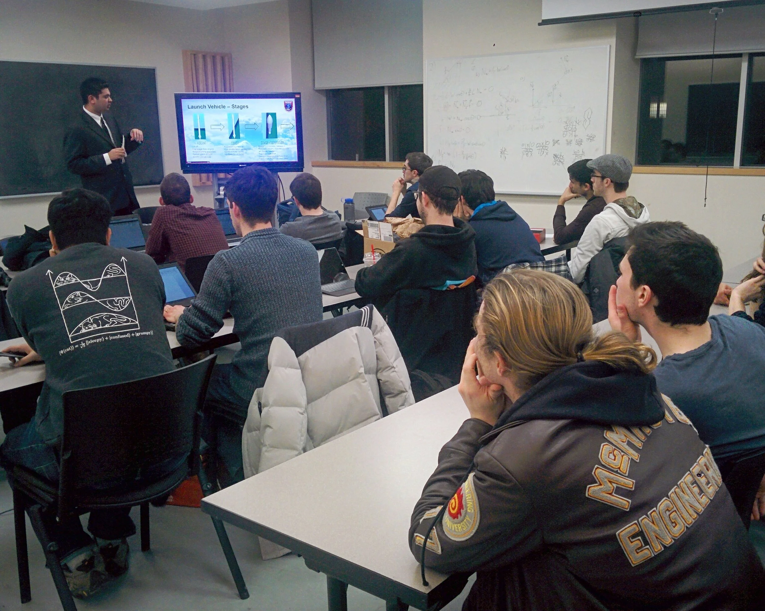 A college classroom with students attentively listening to a male instructor giving a presentation. The instructor stands next to a screen displaying a slide about launch vehicle stages, and some students are using laptops or taking notes.