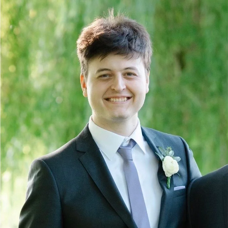 Young man in a tuxedo with a boutonniere, smiling outdoors with a lush green background.