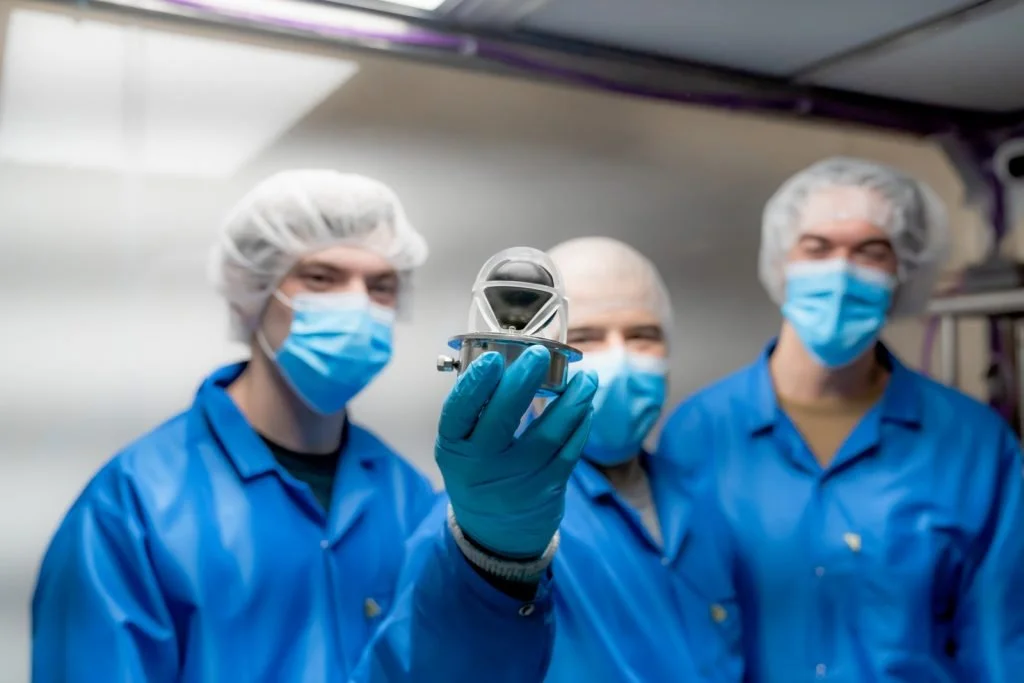 Three scientists in blue lab coats, hairnets, and face masks holding a small metallic object in a laboratory or cleanroom setting.