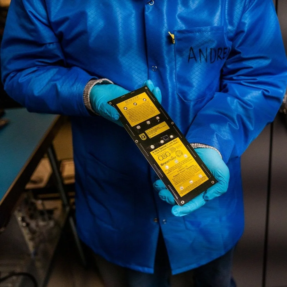 A person wearing a blue jacket with the name 'André' written on it, holding a circuit board or electronic component with yellow labels in an indoor setting.
