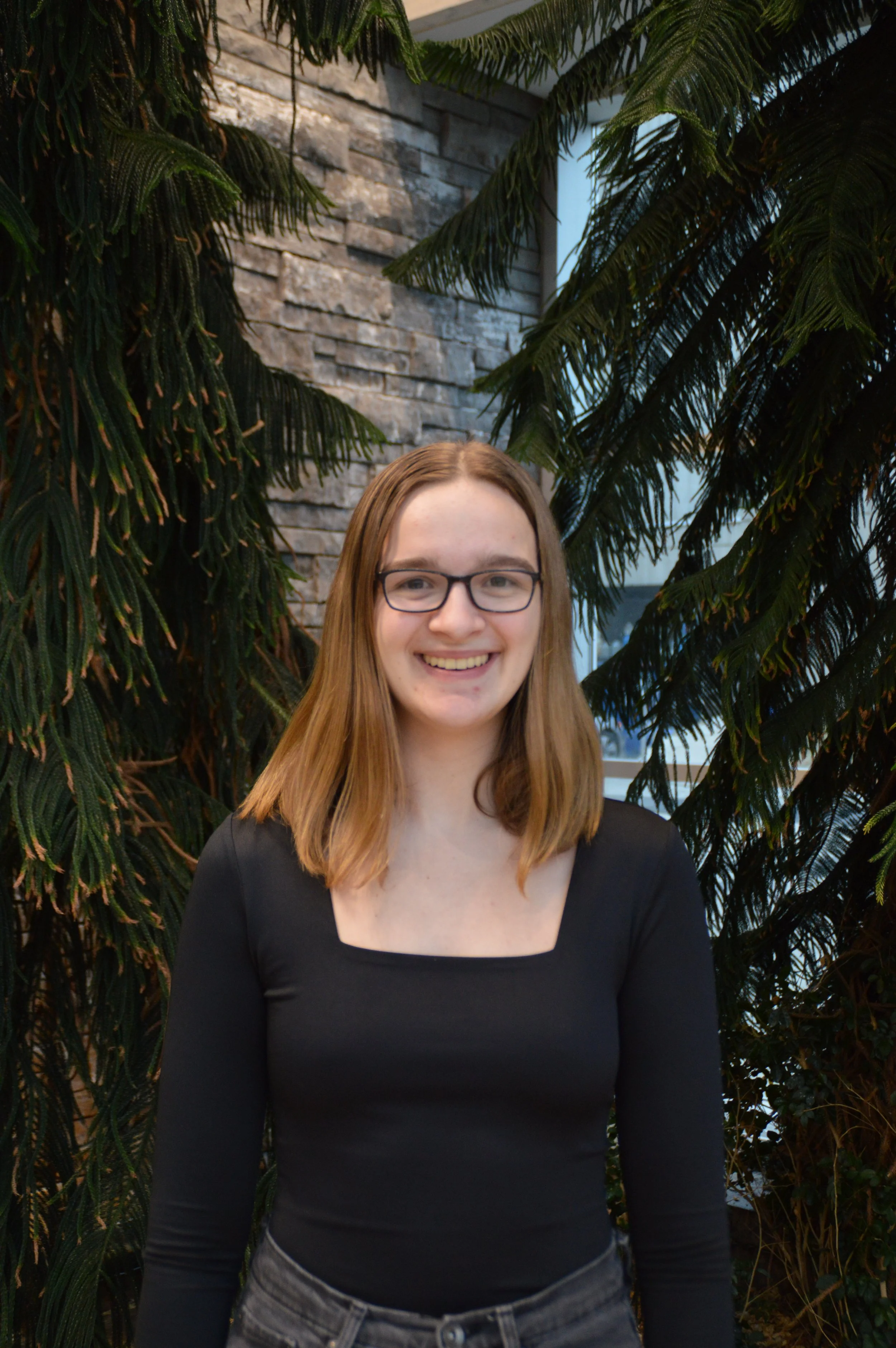 A young woman with shoulder-length red hair, glasses, and a black long-sleeve top, smiling in front of lush green plants and a brick wall interior.
