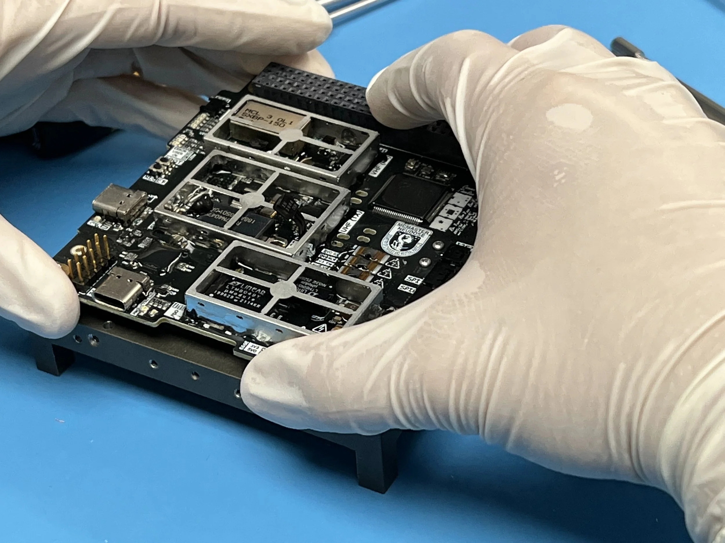 Person wearing white gloves handling a circuit board with multiple electronic components on a blue surface.